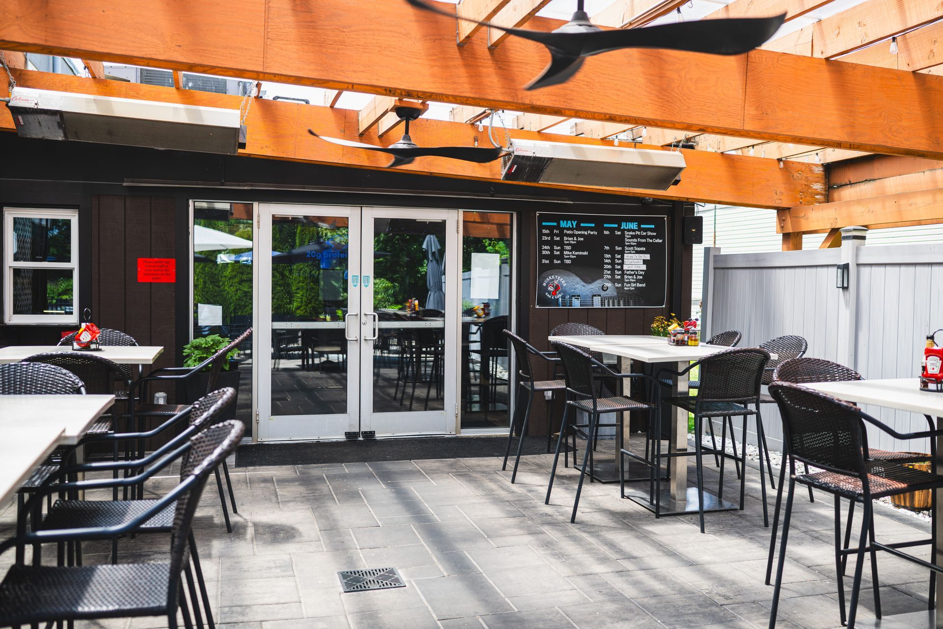 Outdoor patio dining area with black tables, chairs, and a wooden pergola in front of glass doors.