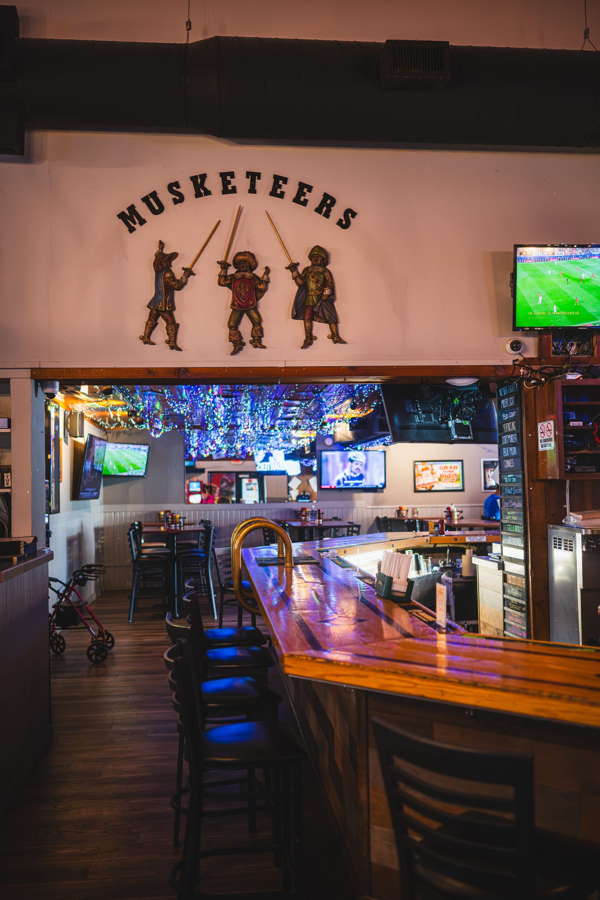 Dimly lit bar interior with a curved wooden counter, high stools, TVs, and a wall sign reading “Musketeers.”