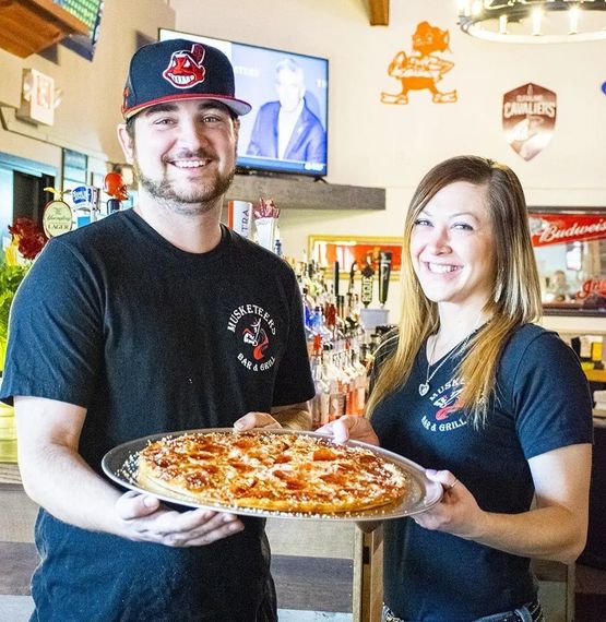 Two smiling employees holding a pizza inside a pizzeria