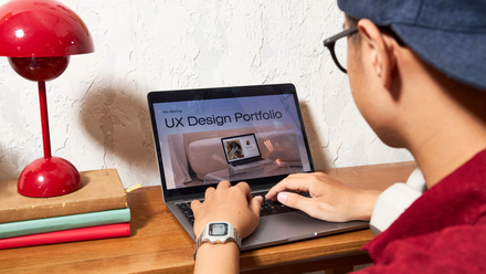 Person working on a laptop, viewing a UX design portfolio. Red lamp and stacked books sit nearby.