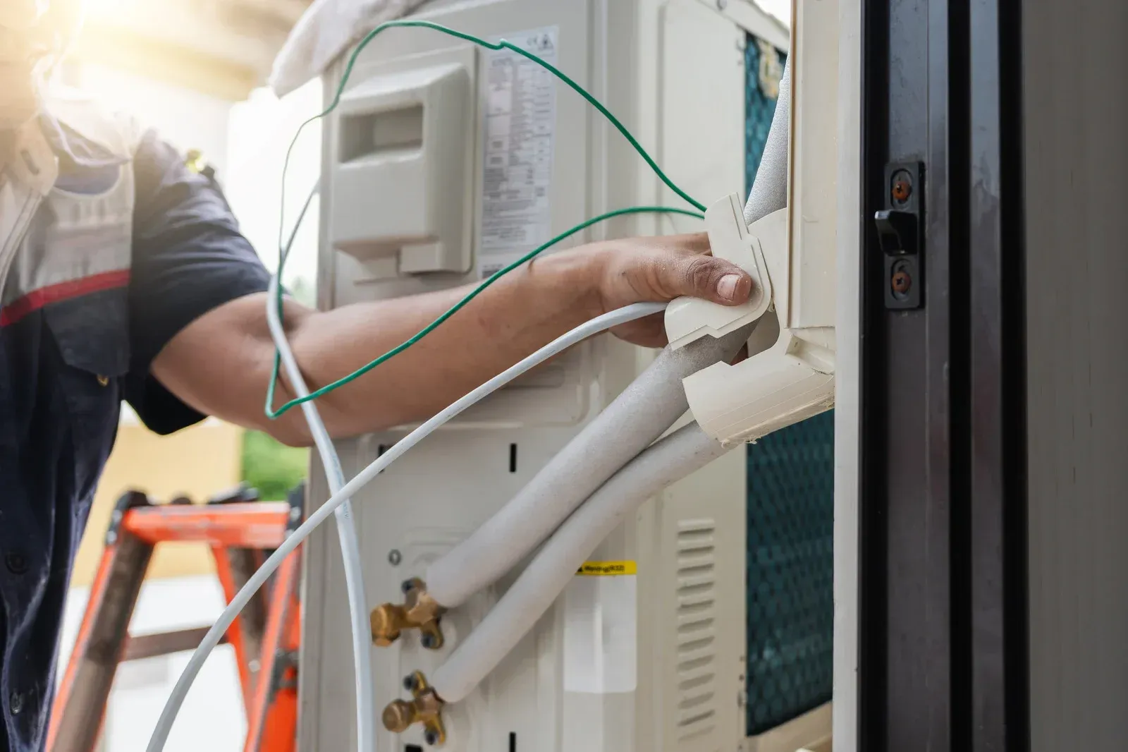 Person installs AC unit; outdoors, next to door. Wires and tubing connected to unit; ladder in background.