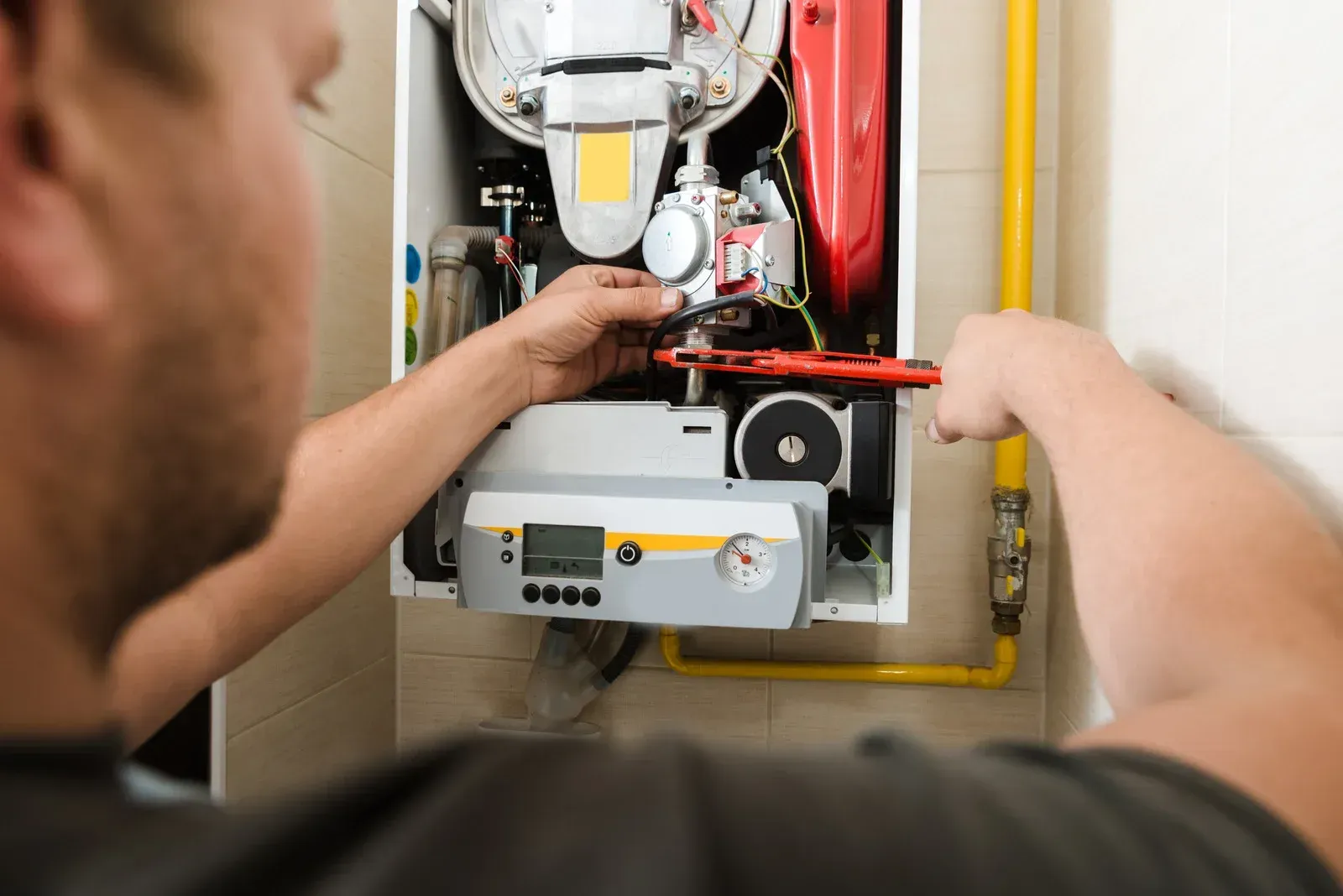 Person working on a heating system with tools. White cabinet, yellow pipe, and gray control panel are visible.