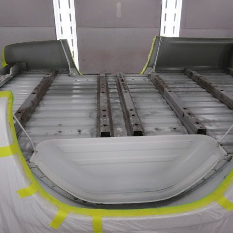 Interior of a vehicle being painted, showing gray metal frame and taped sections, in a white workshop.