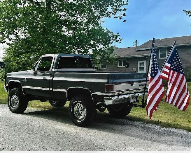 Dark truck with American flags parked on a road, house and trees in the background.