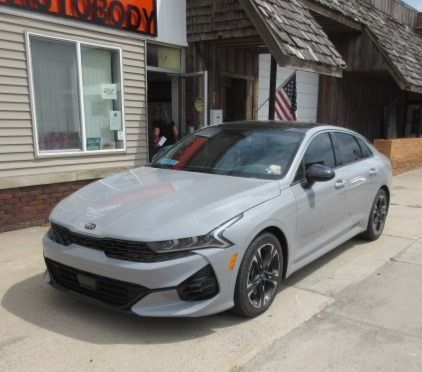 Gray Kia K5 sedan parked in front of an auto body shop with a wooden exterior and American flag.