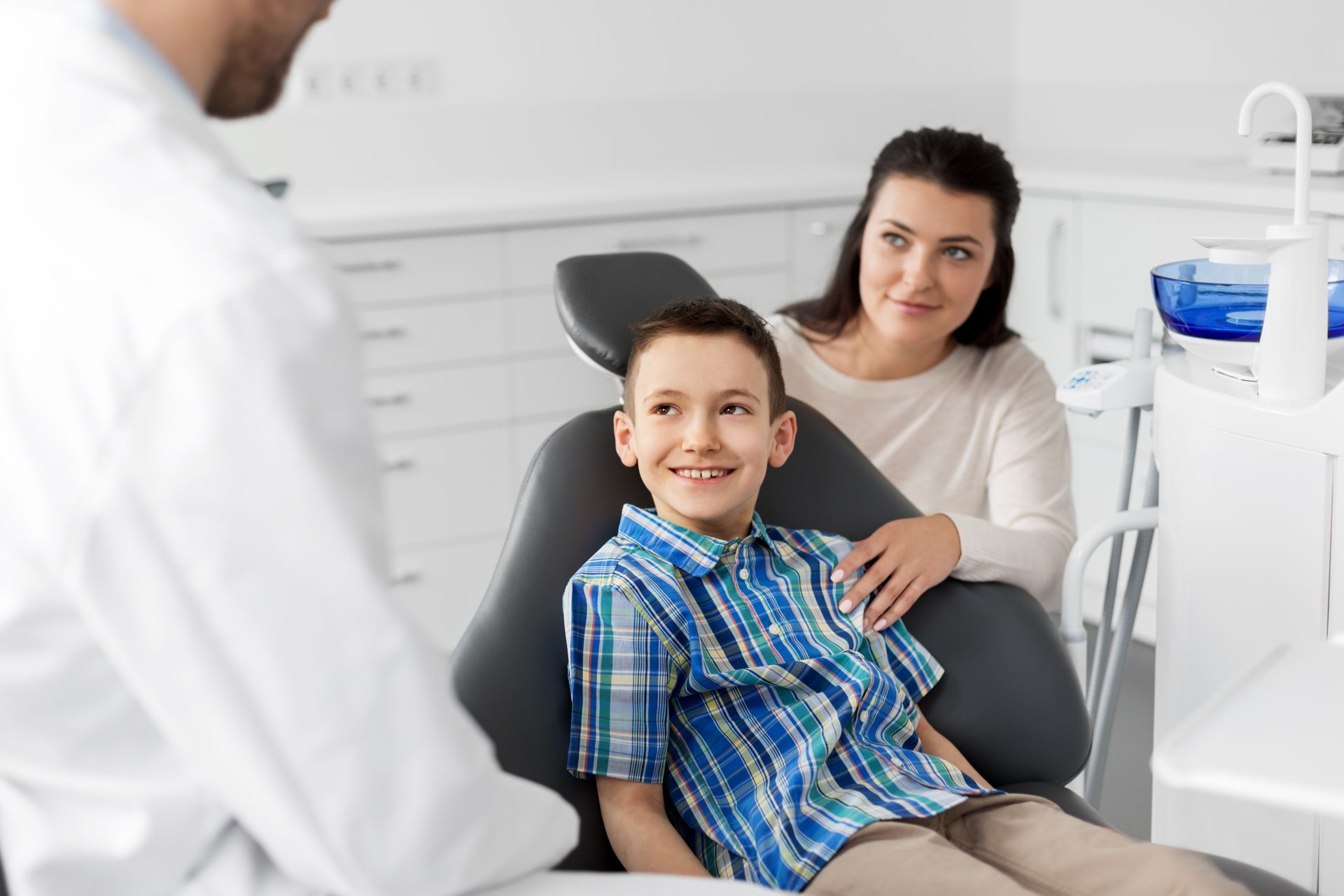 A BOY IN A DENTIST’S CHAIR, SMILING AND LOOKING AT A DENTIST, WHILE HIS MOTHER STANDS BESIDES HIM.