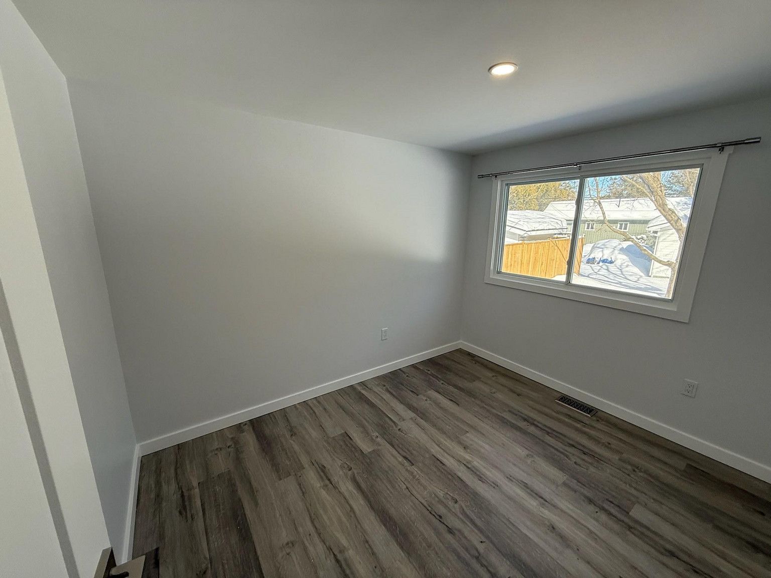 An empty room with light gray walls, dark wood-look flooring, and a window with a white frame.