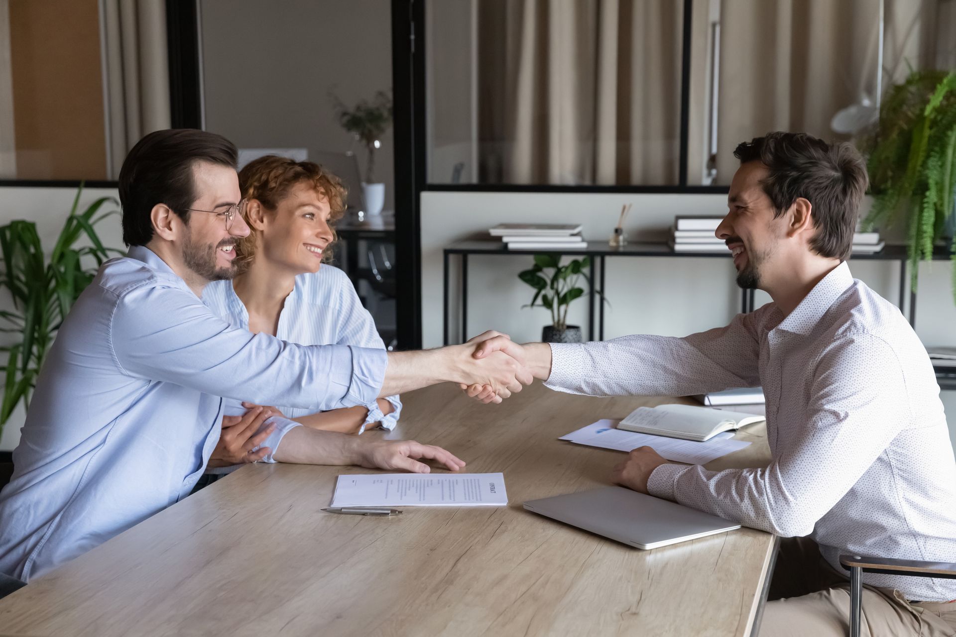 Three people in a meeting room shaking hands across a table, with documents and a laptop nearby.