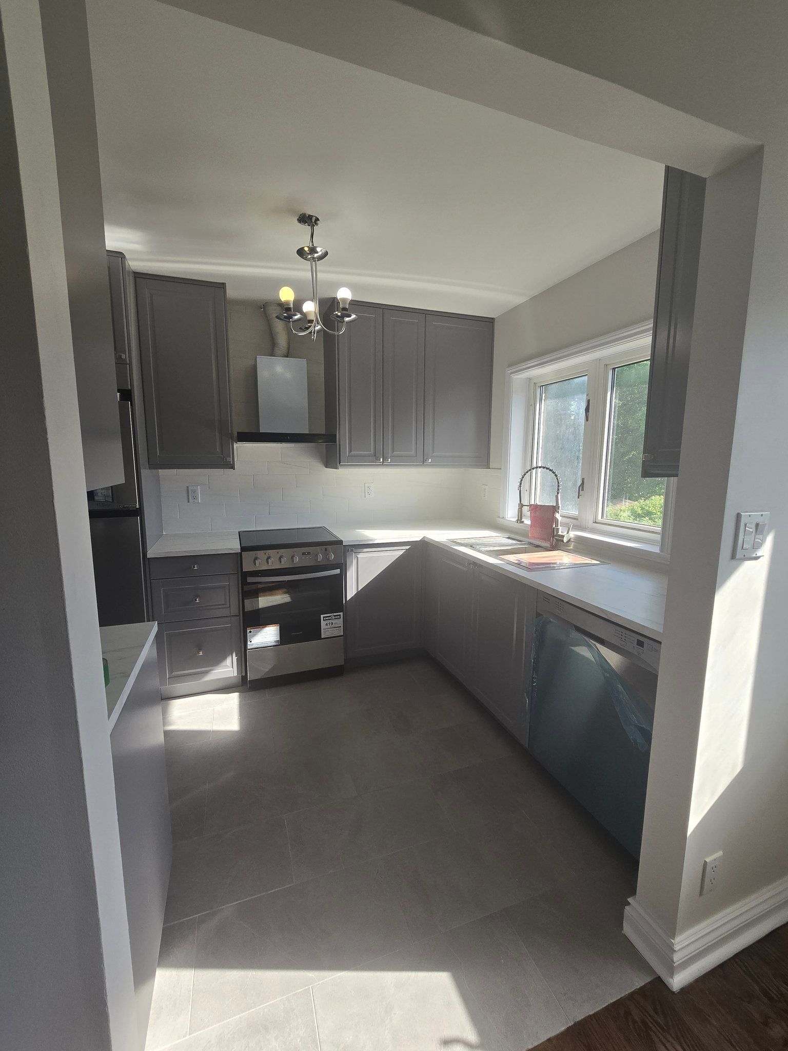 A kitchen with gray cabinets , a stove , a dishwasher and a window.
