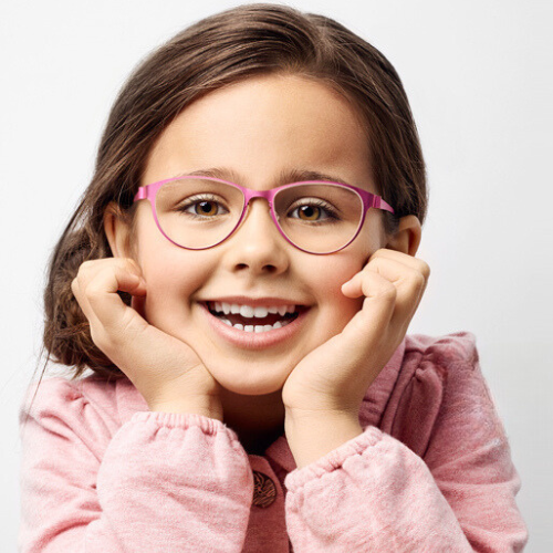 Girl with pink glasses, smiling broadly, resting chin on hands.