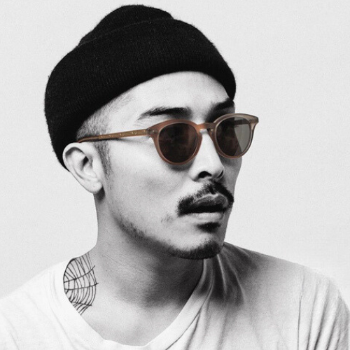 Man wearing sunglasses and a black beanie, looking to the side. He has a mustache and a neck tattoo.