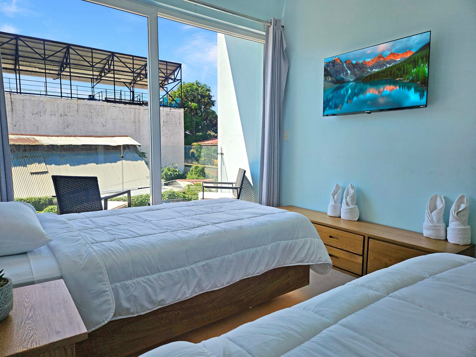 Bedroom with two beds, large window, TV, light blue wall. Outside view of a structure.