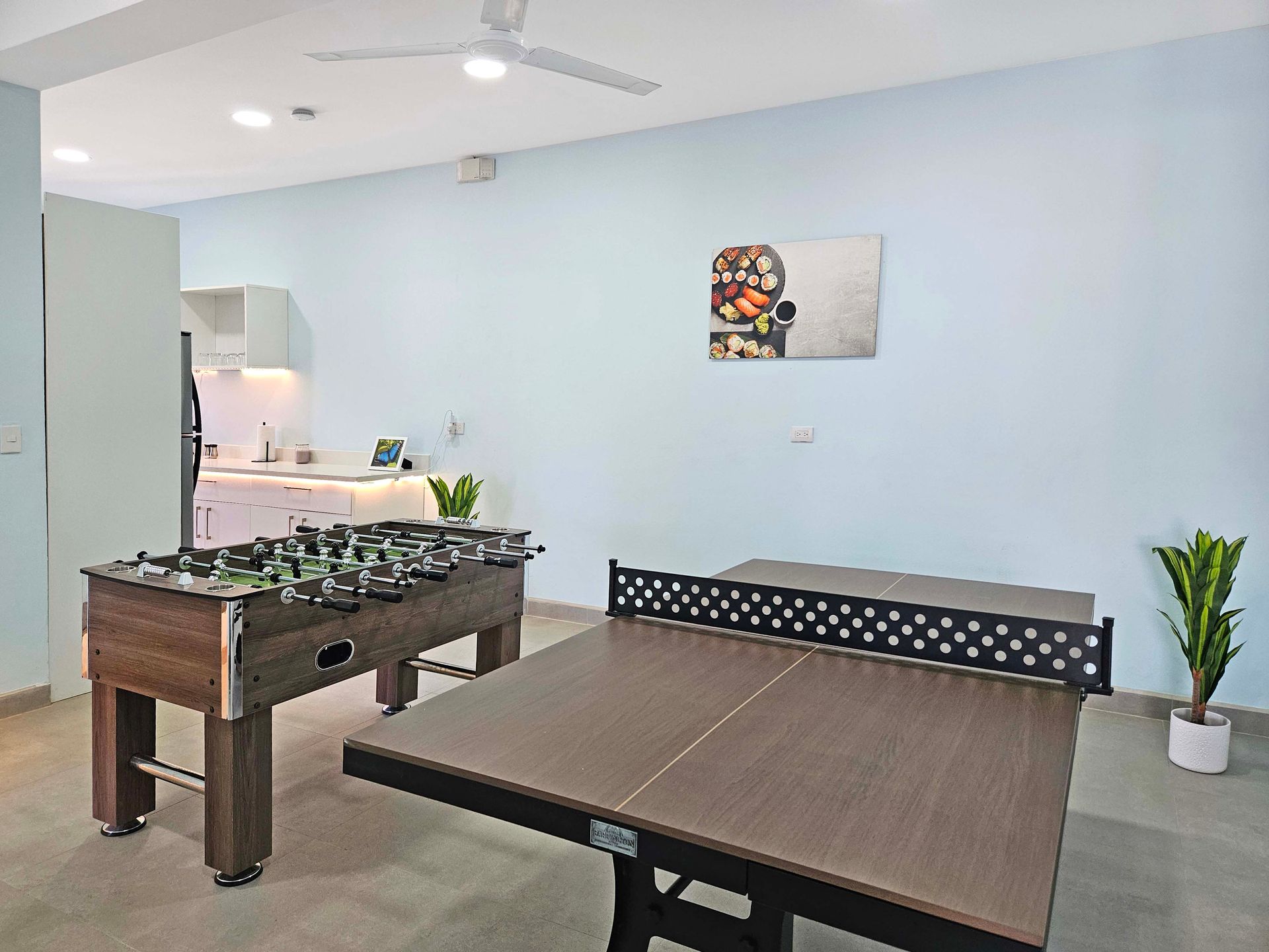 Game room with foosball and ping pong tables; light blue walls, plants, and a small kitchen area.
