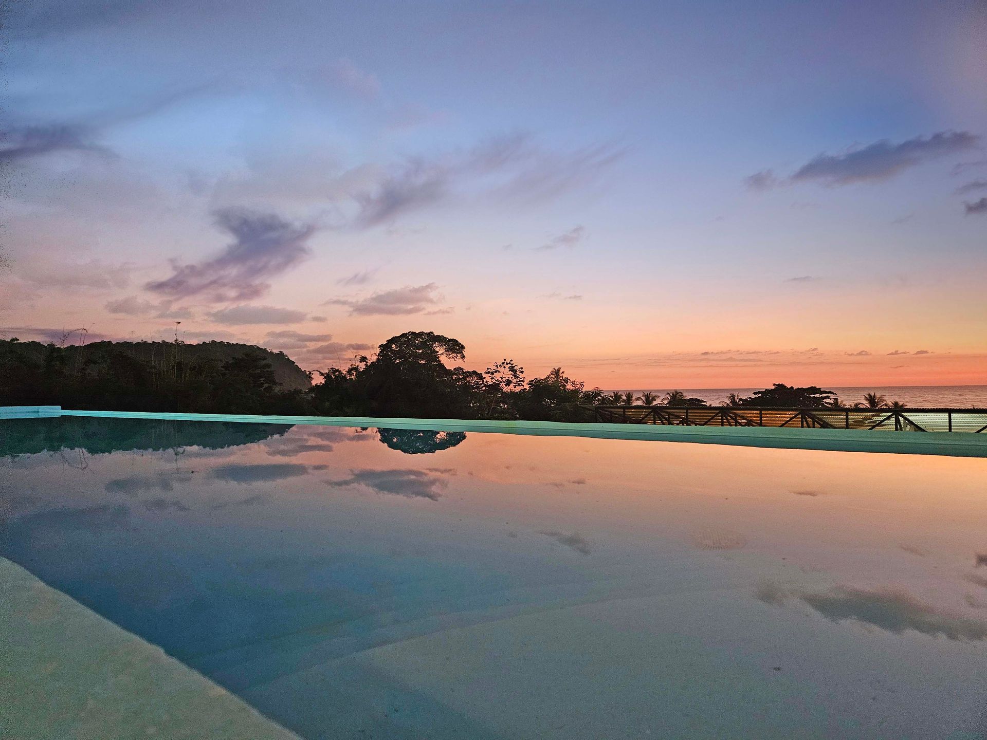 Sunset over a pool, reflecting the colorful sky, with a dark tree line on the horizon.