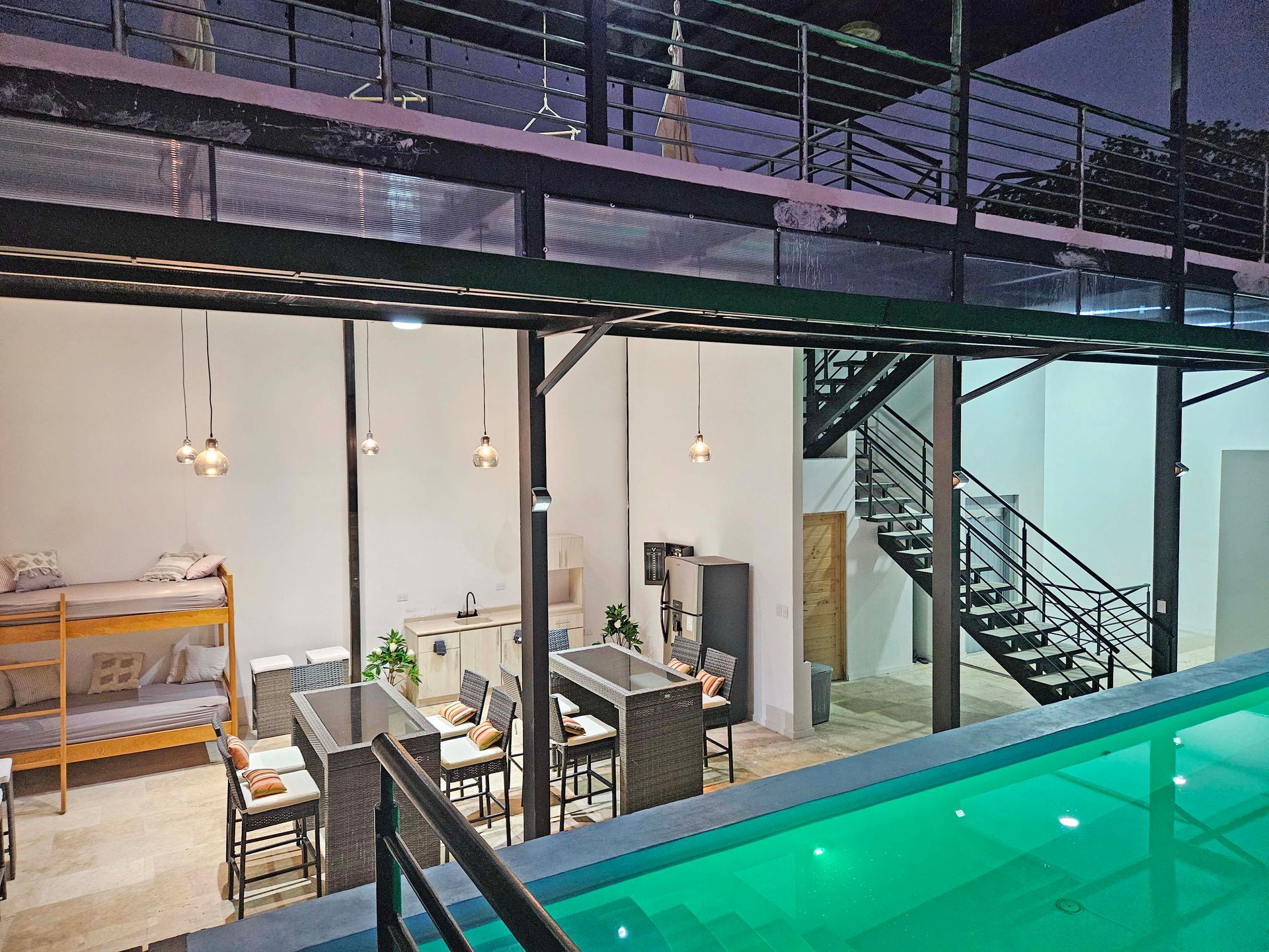 Poolside lodging: bunk beds, dining tables, metal staircase, and pool with green water under a mezzanine.