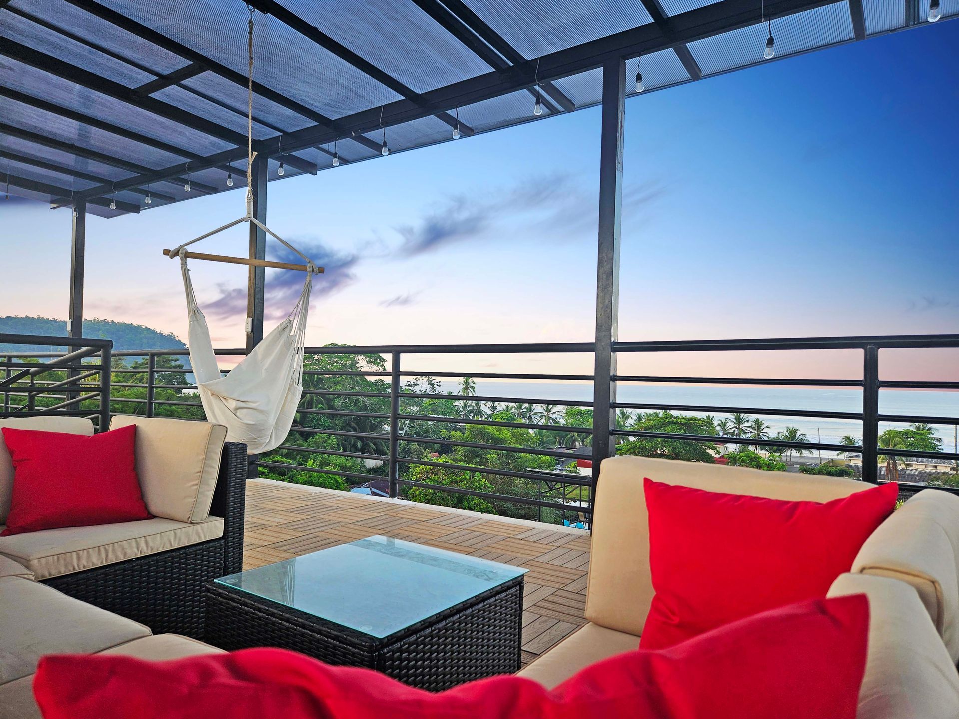 Rooftop patio with seating, hammock, and ocean view at sunset. Red pillows, black table, and blue sky.