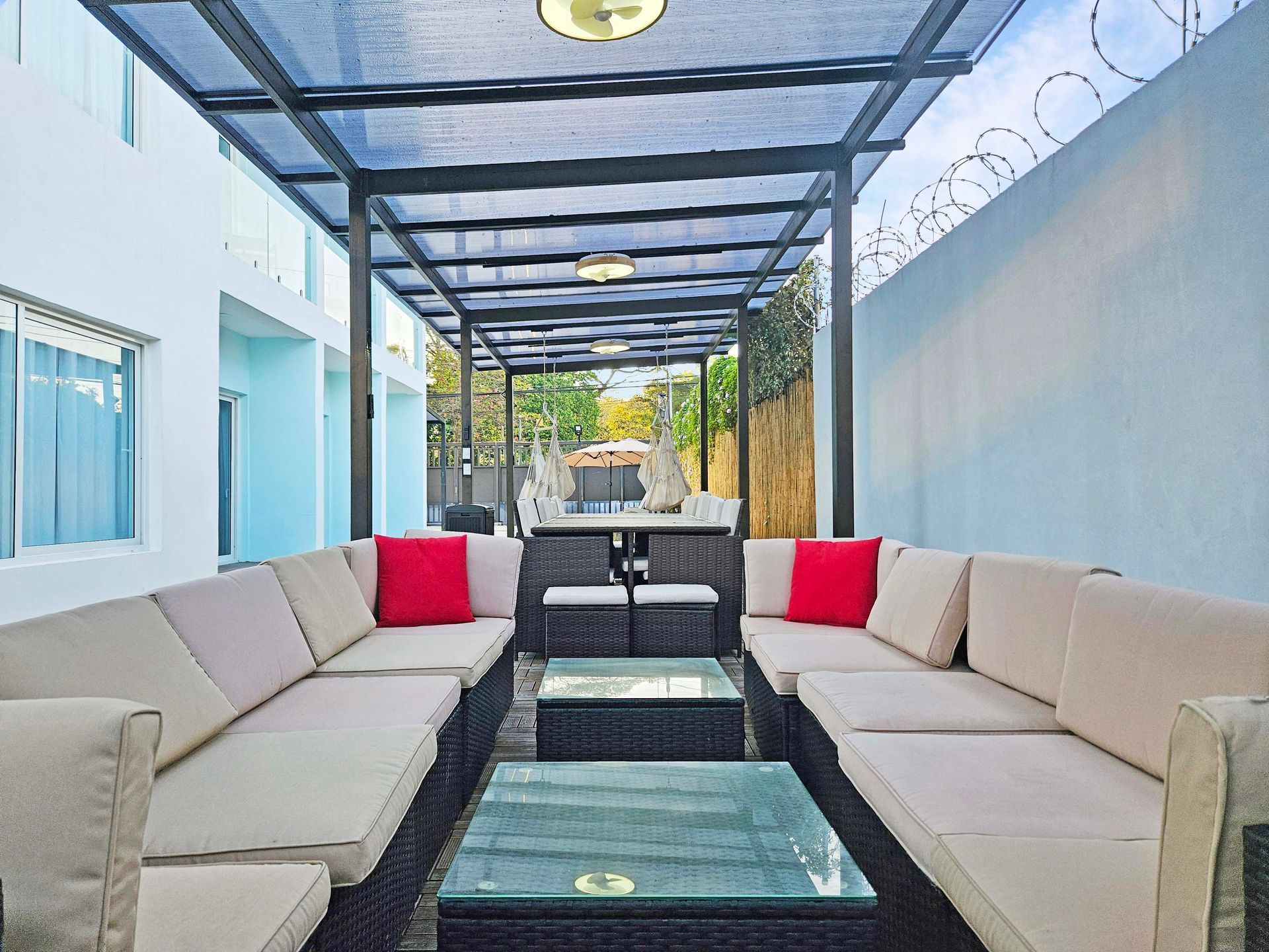 Outdoor lounge area with beige couches, red pillows, and glass coffee tables under a shaded structure.