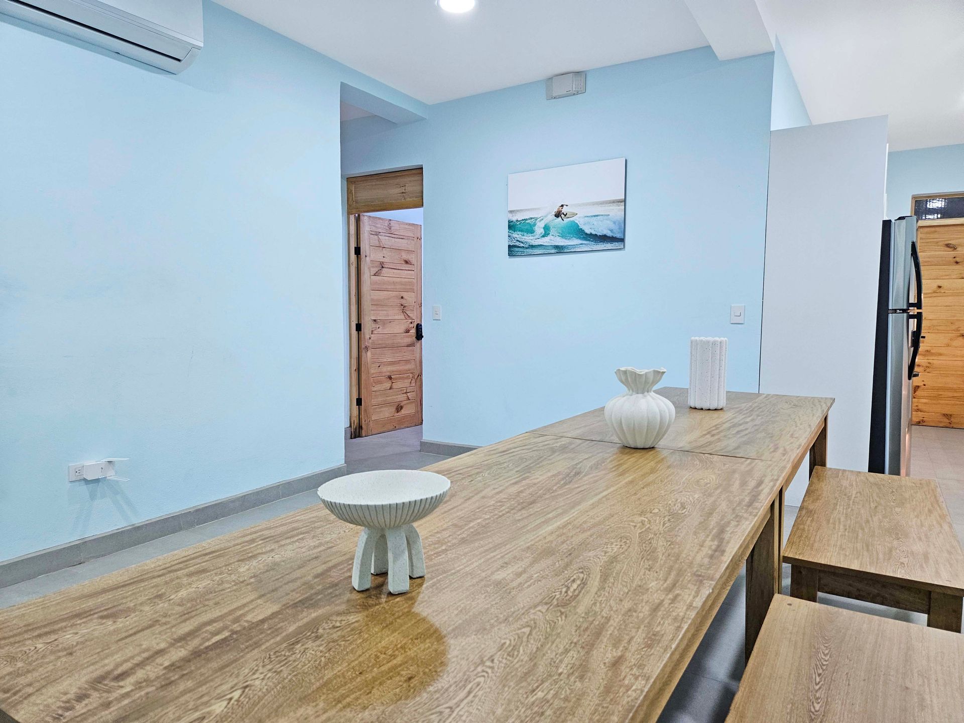 Dining area with long wooden table, benches, light blue walls, art, doorway, and air conditioning unit.