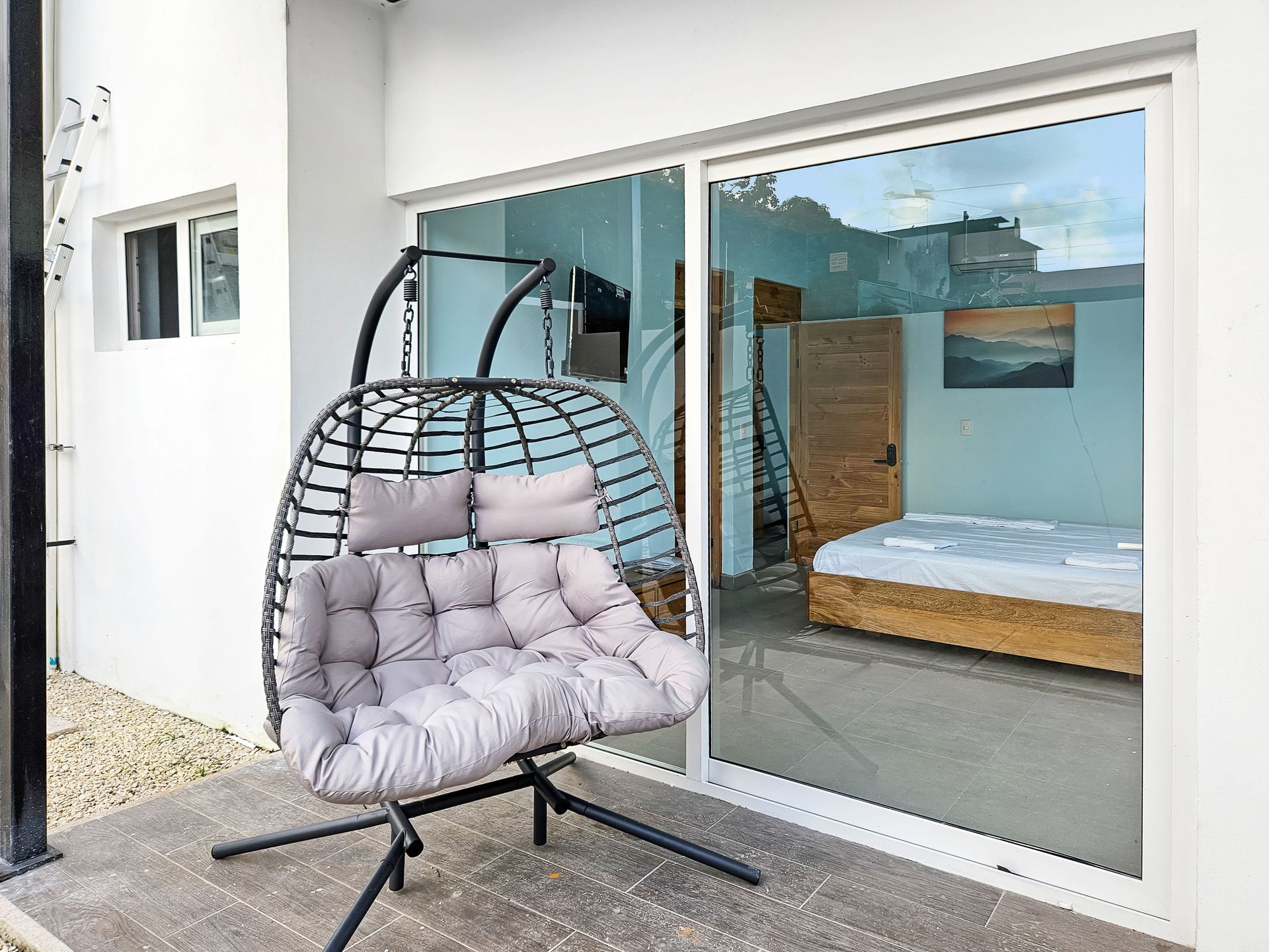 A gray wicker hanging chair on a patio. Sliding glass door shows bedroom with bed.