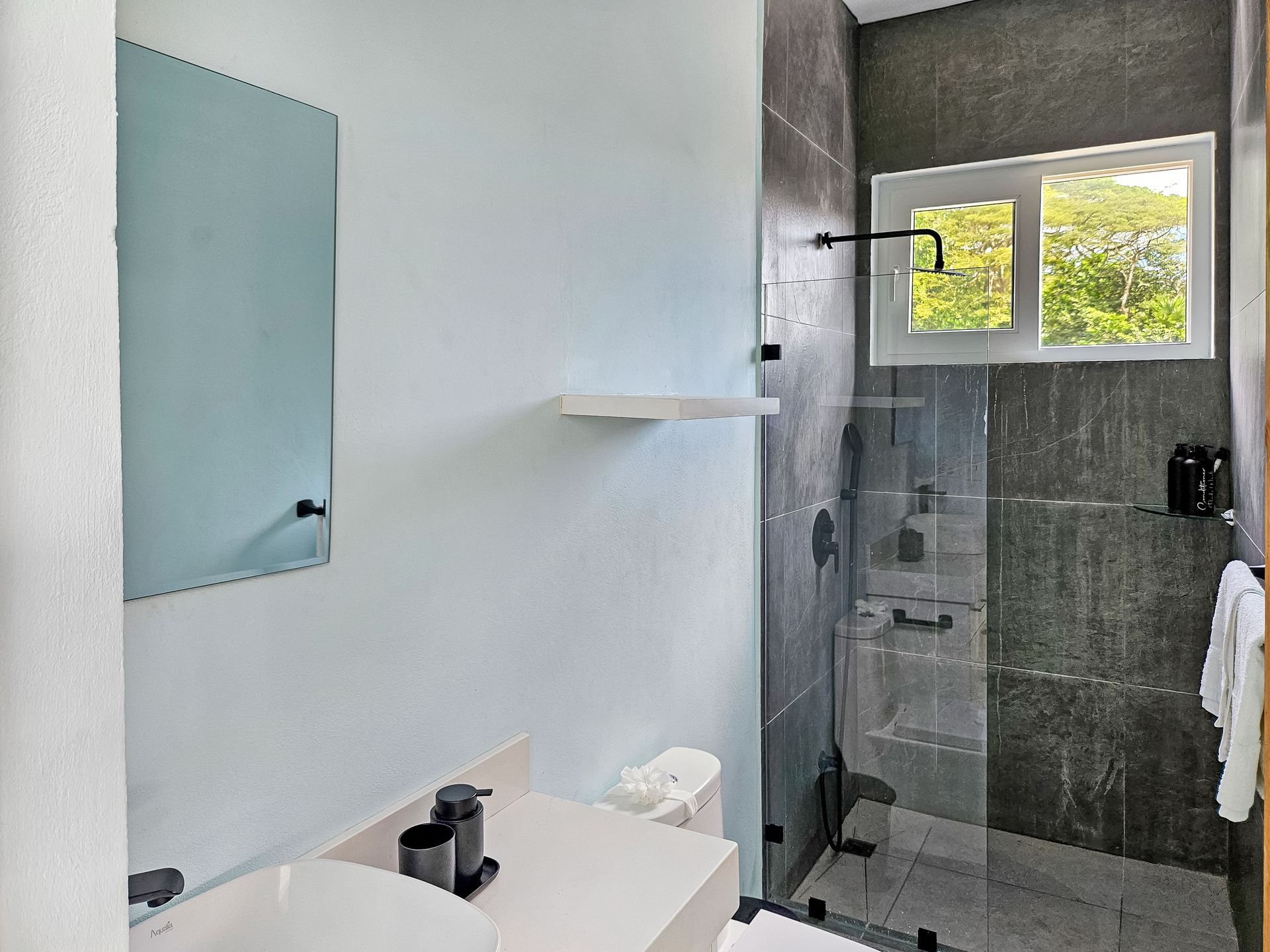 Modern bathroom with white and gray walls, glass shower, and small window looking at trees.