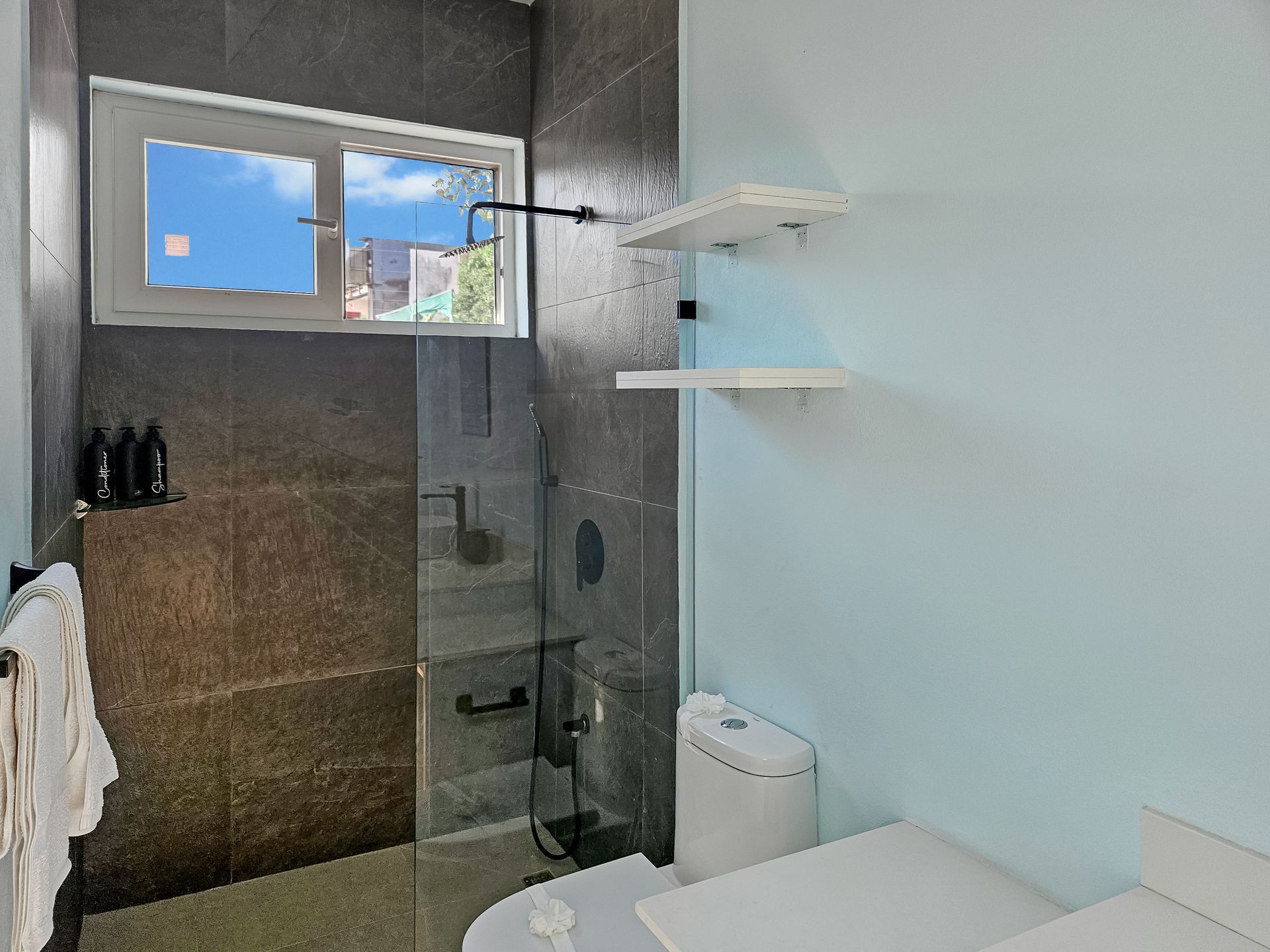 Bathroom with shower behind glass door, toilet, window, light blue and gray walls, two shelves.
