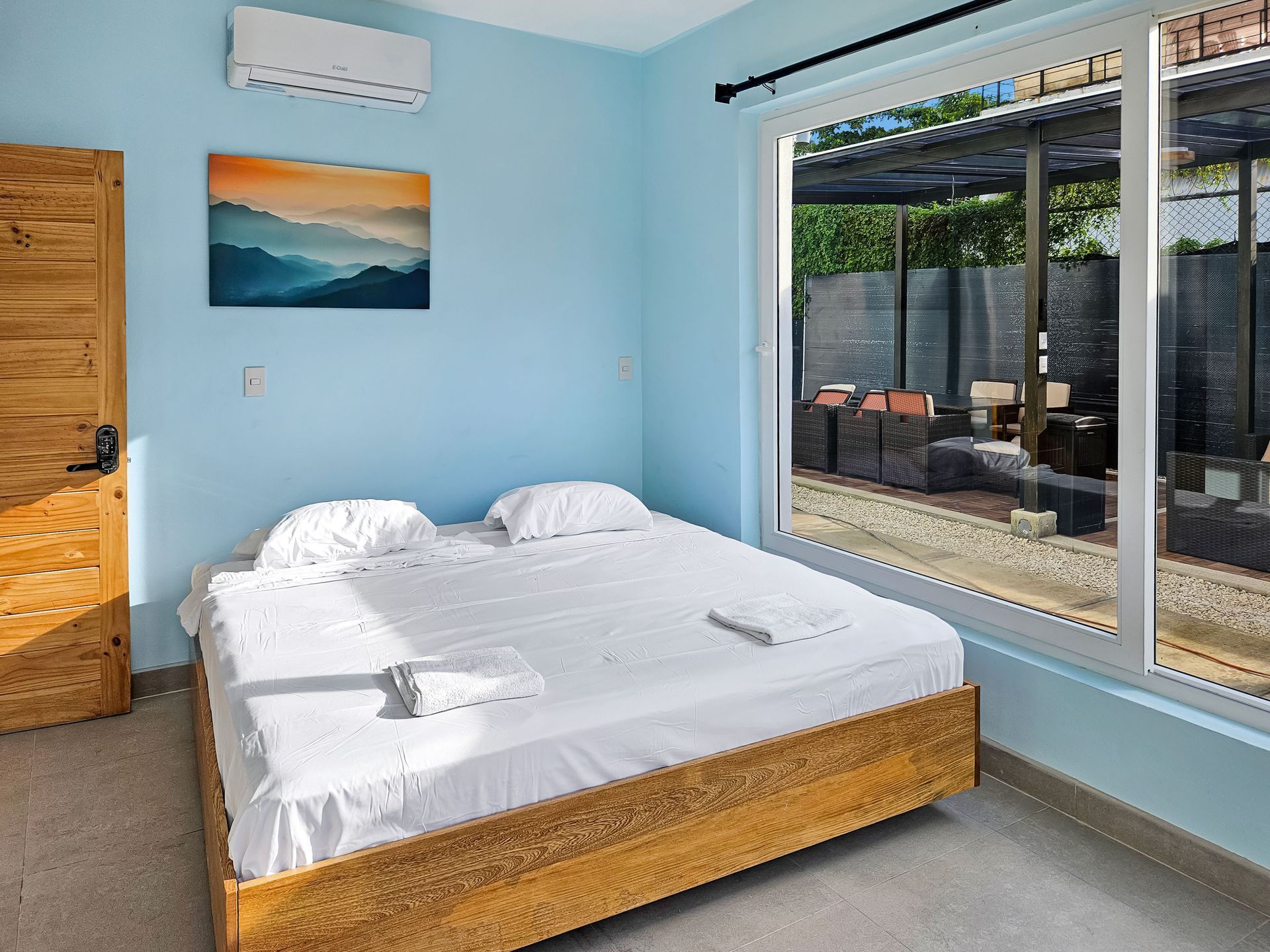 Bedroom with a wooden bed, blue walls, a painting, and a large window overlooking an outdoor area.