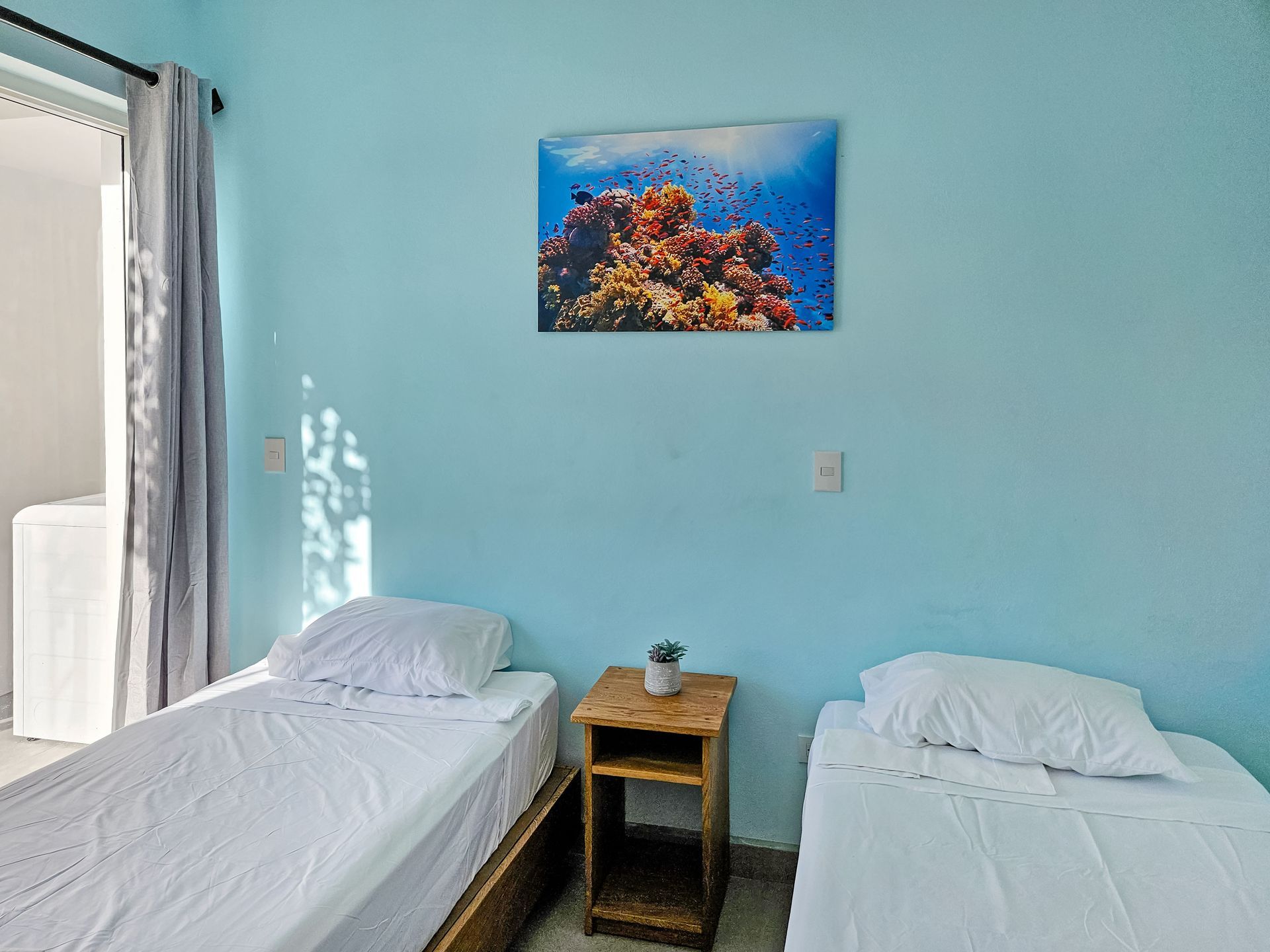 Two twin beds in a light blue room with coral reef art above a small wooden nightstand.
