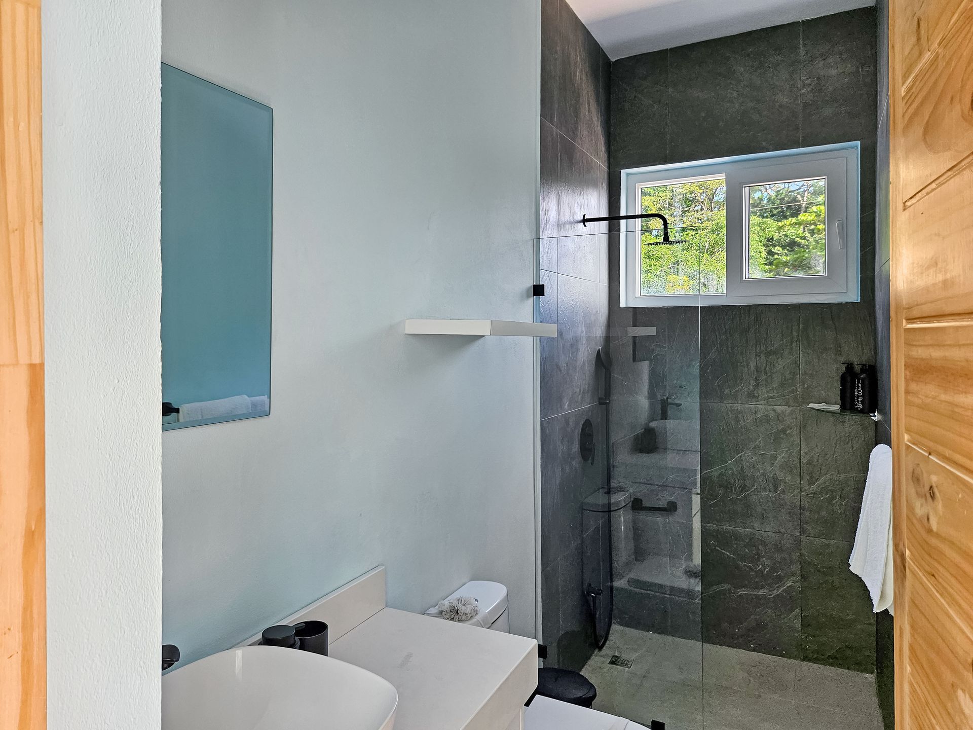 Bathroom with gray tiled shower, white sink, and wooden accents, with a window.