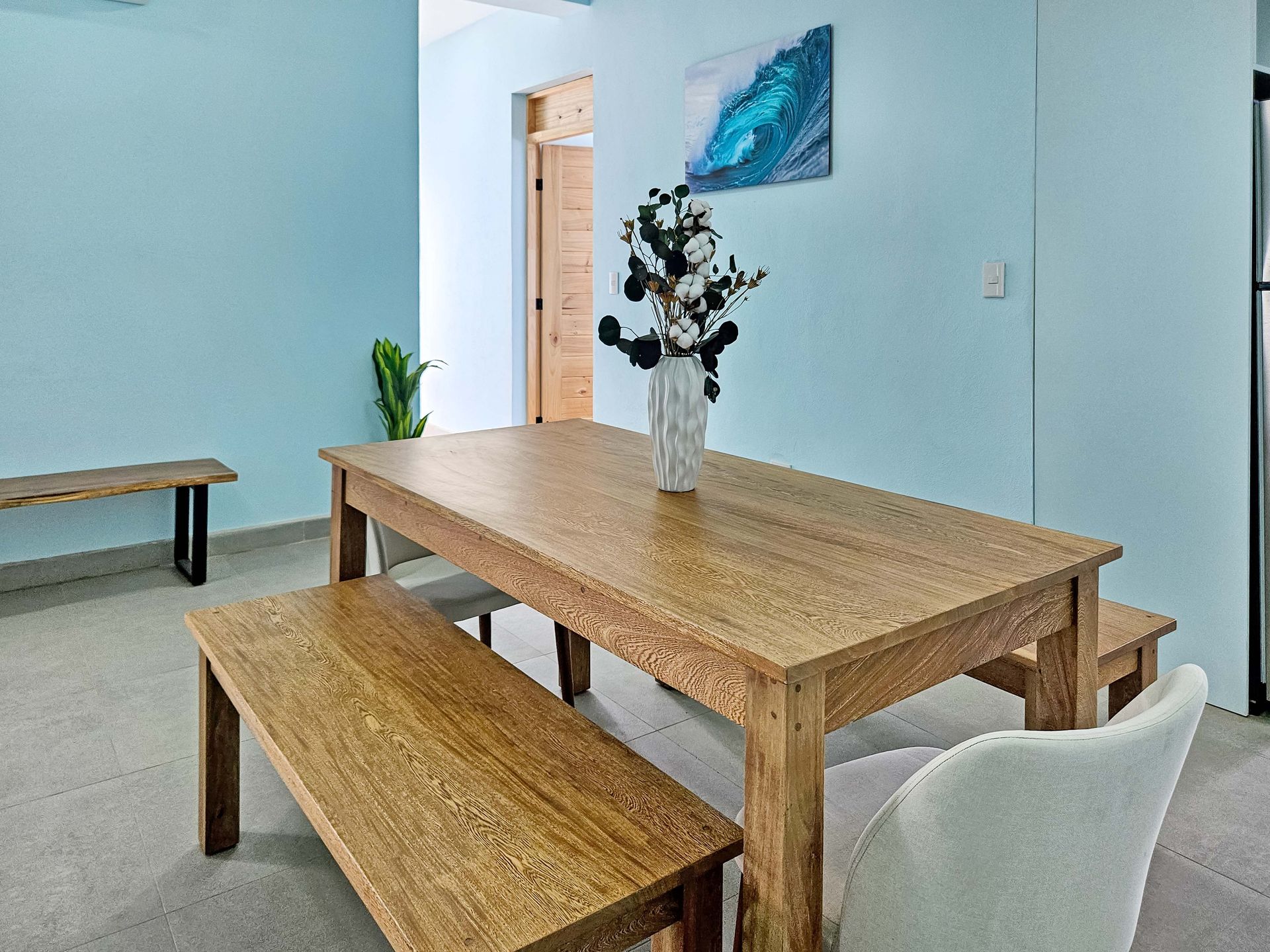 Wooden dining table with benches in a light blue room; a vase of flowers is centered on the table.