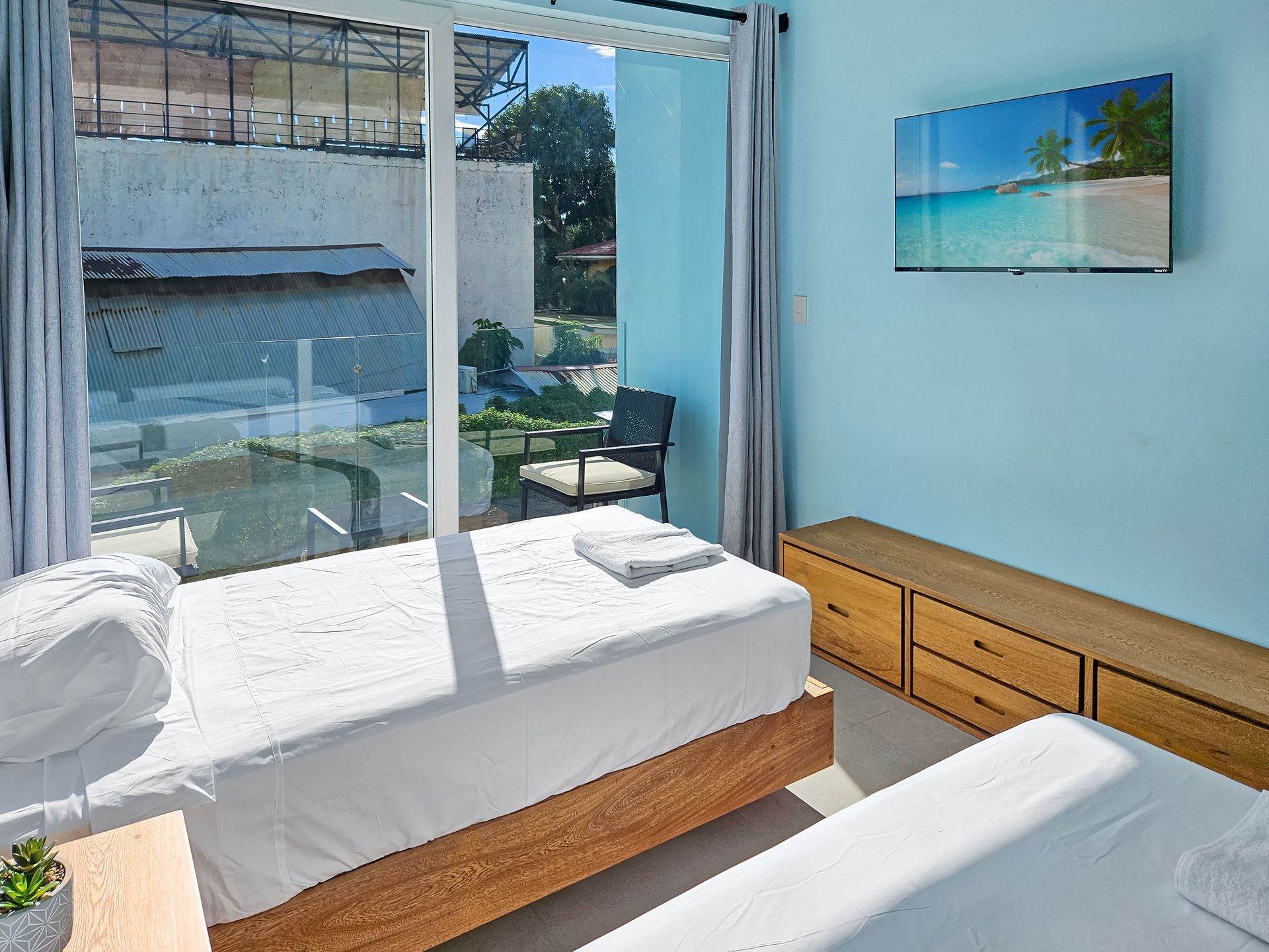 Bedroom with two white beds, blue walls, wooden furniture, and a window overlooking a small garden.