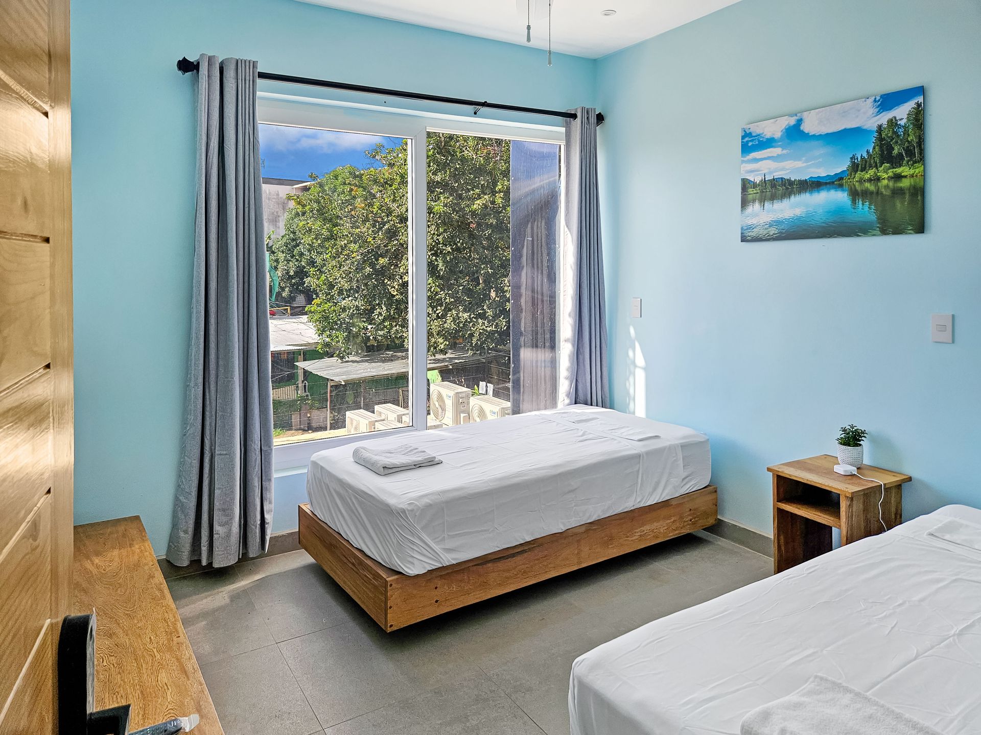 Bedroom with two twin beds, large window, light blue walls, art, and a nightstand.