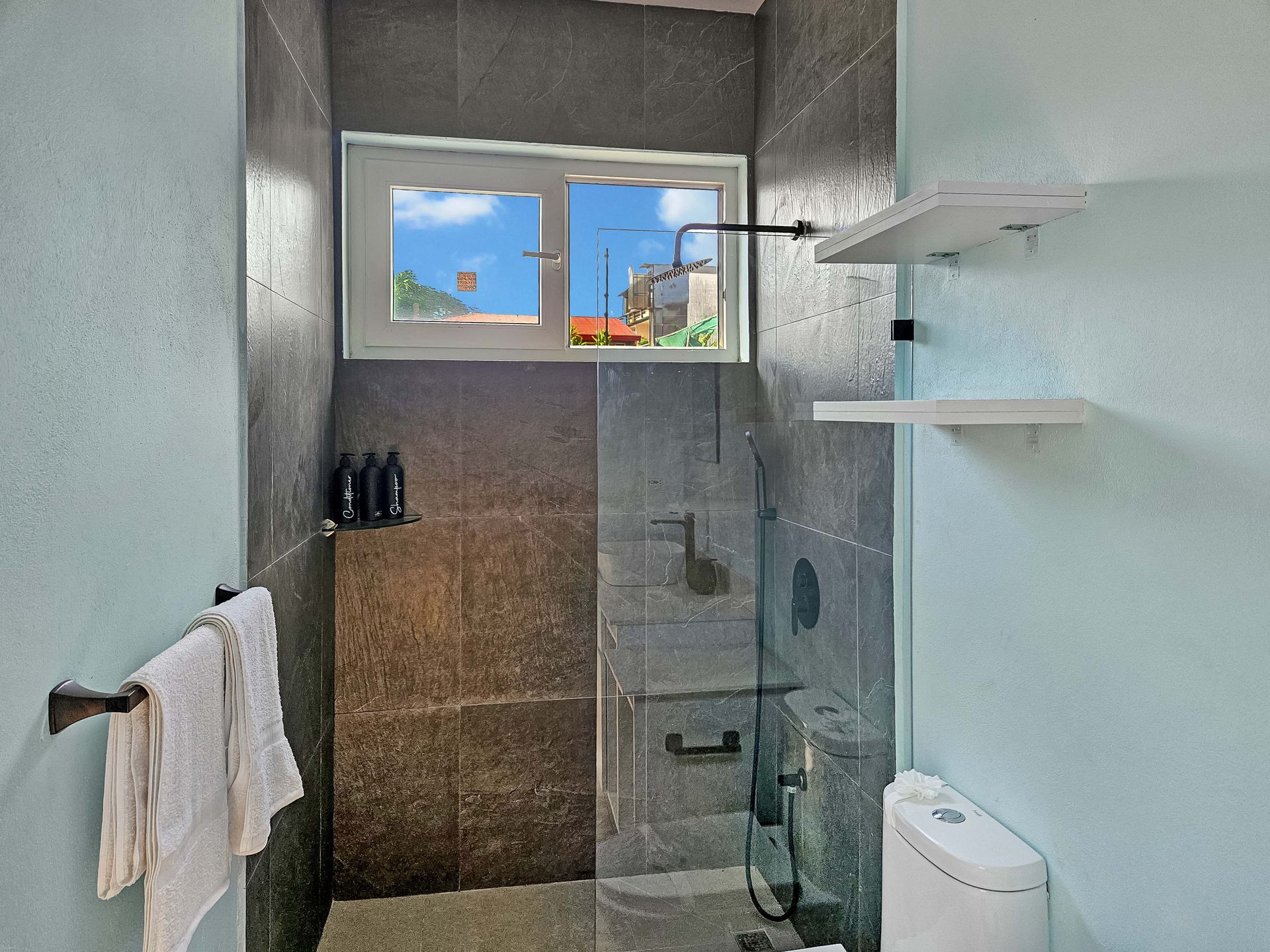 Bathroom with gray tiled shower, window, white shelves, toilet, and towel rack with white towels.