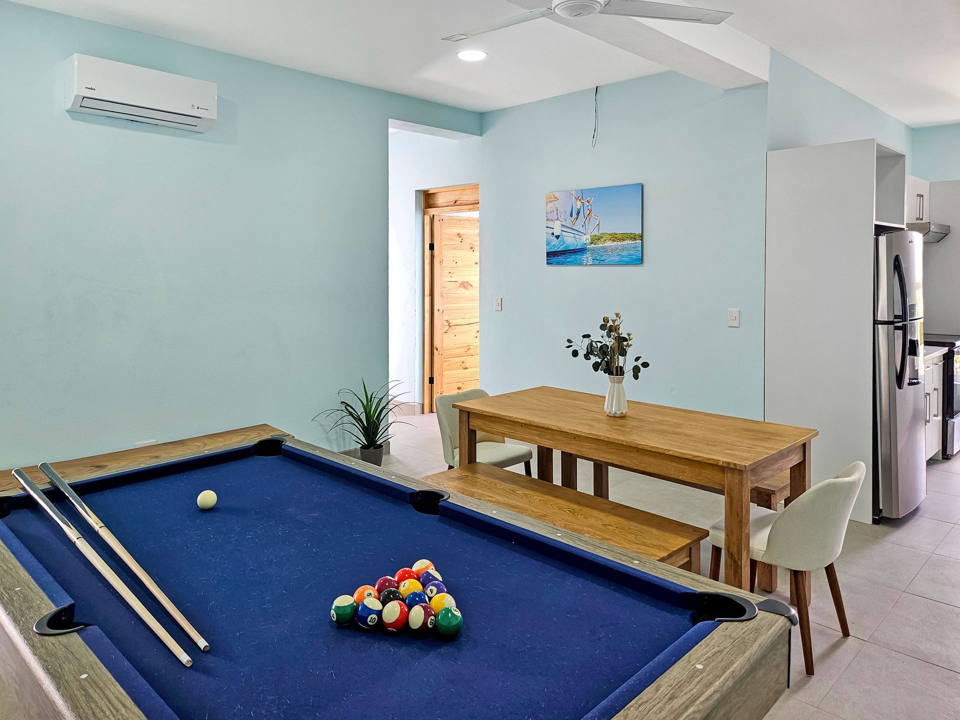 Pool table in a room with a dining table and kitchen. Light blue walls, wood door, and a painting.