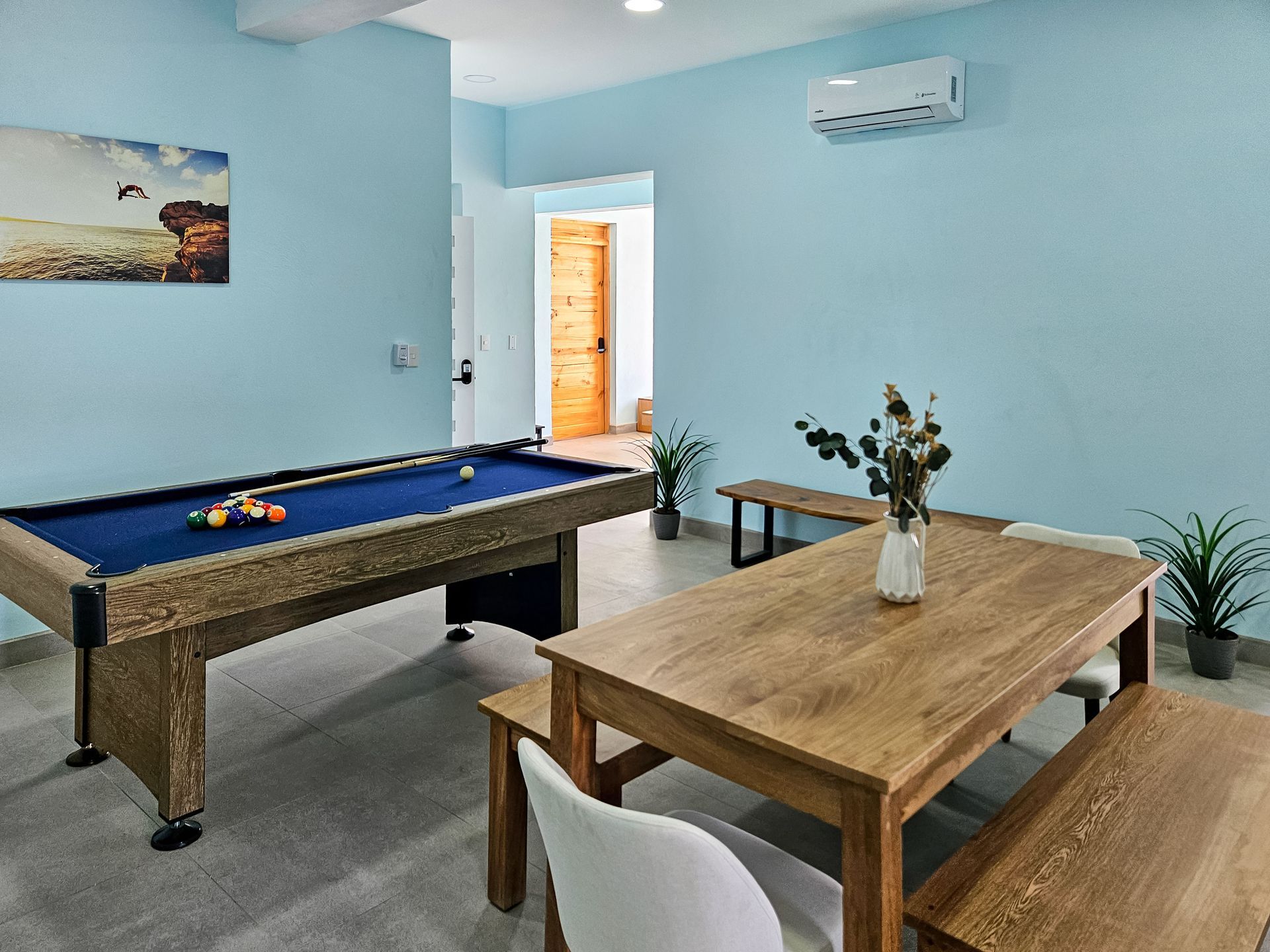 Pool table and dining table in a light blue room with plants and a painting.