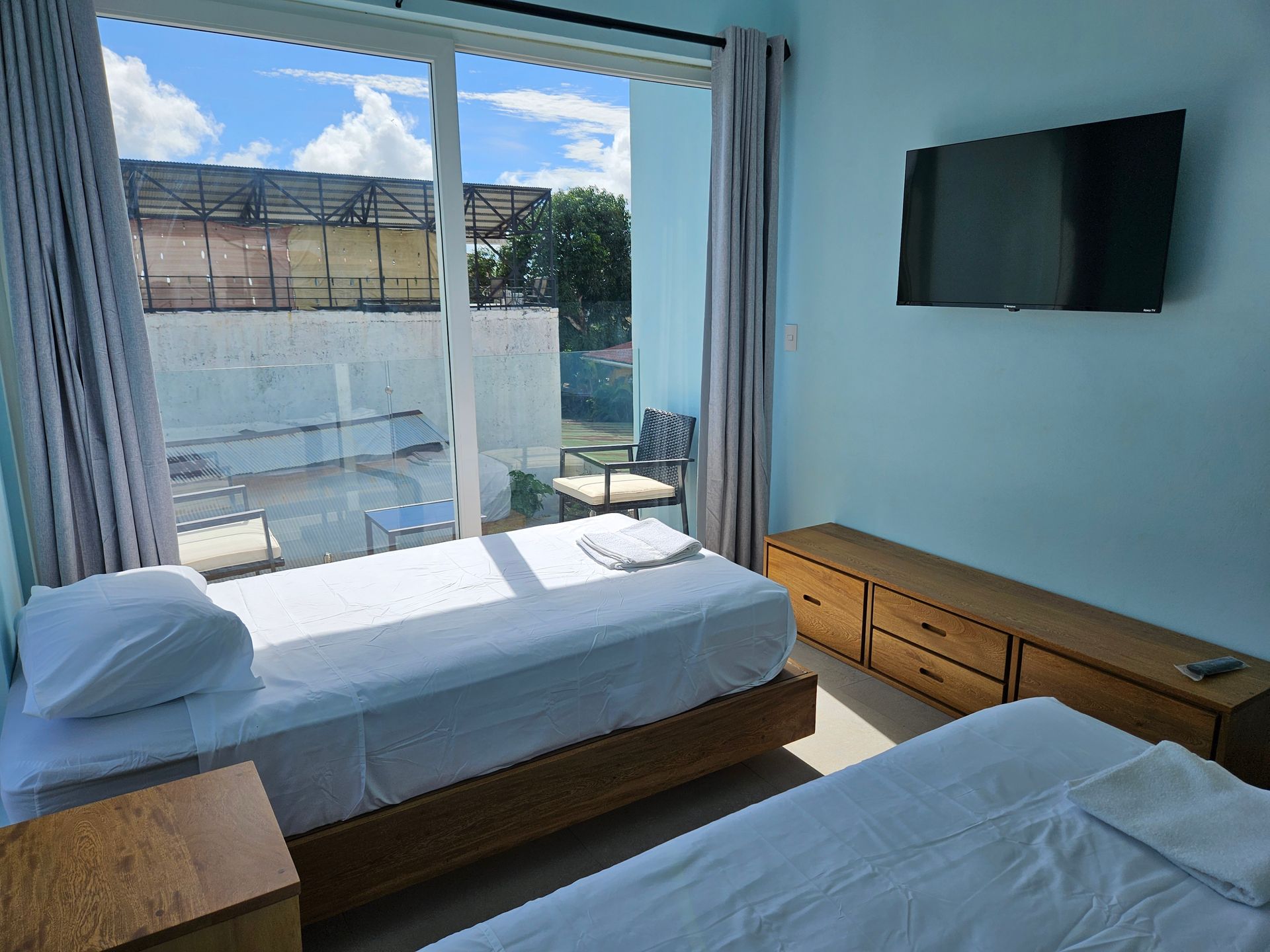 A bright hotel room with two twin beds, a balcony, and a wall-mounted TV.
