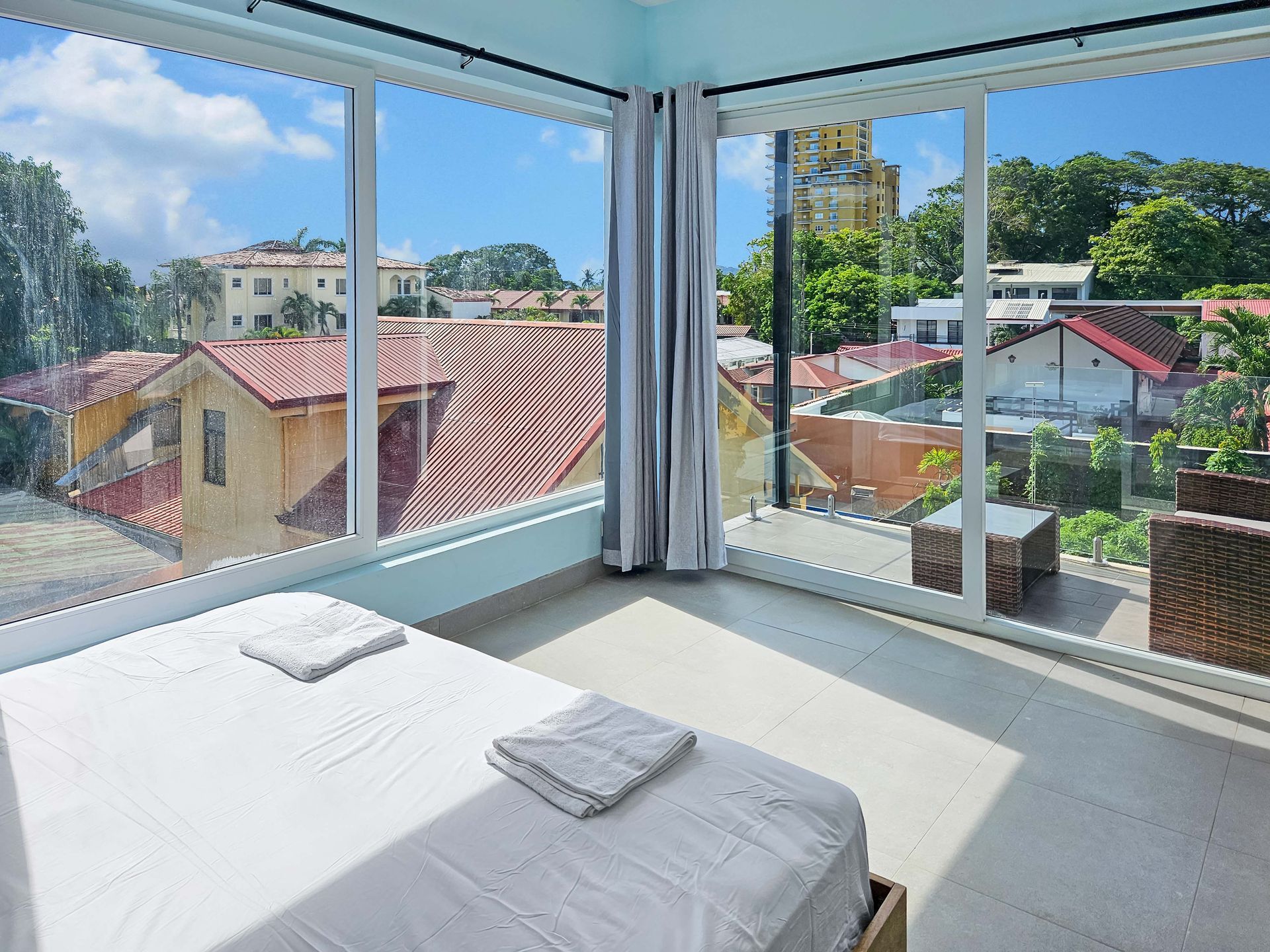 Bedroom with large windows, city view, bed with towels, light blue walls, curtains.