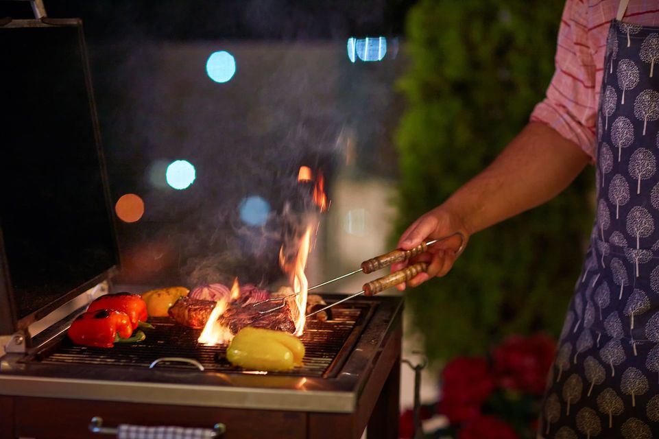 Person grilling food on a barbecue at night; flames visible; colorful lights in background.
