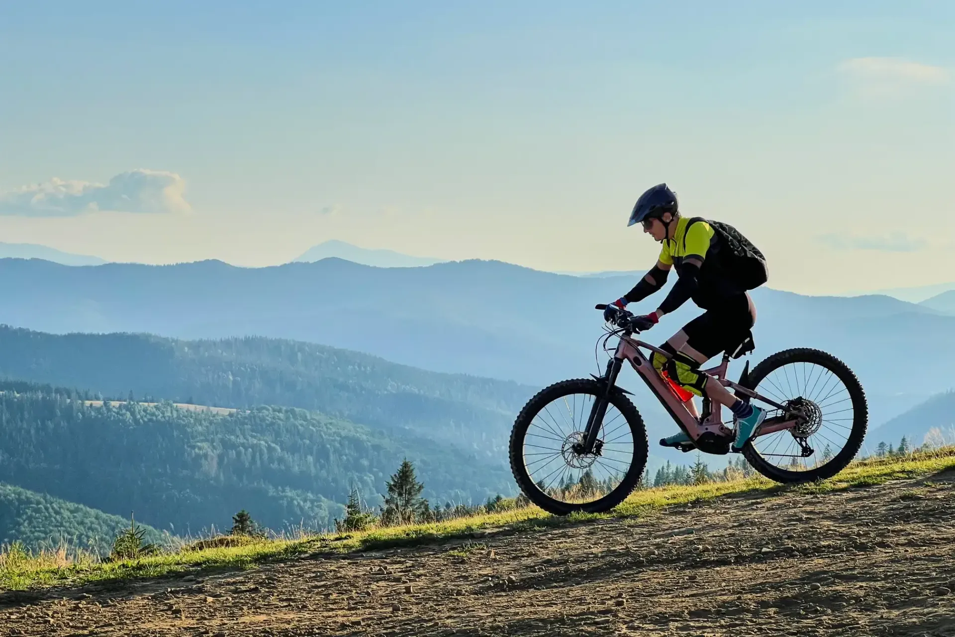 Mountain biker rides uphill on a dirt path with mountain range backdrop.