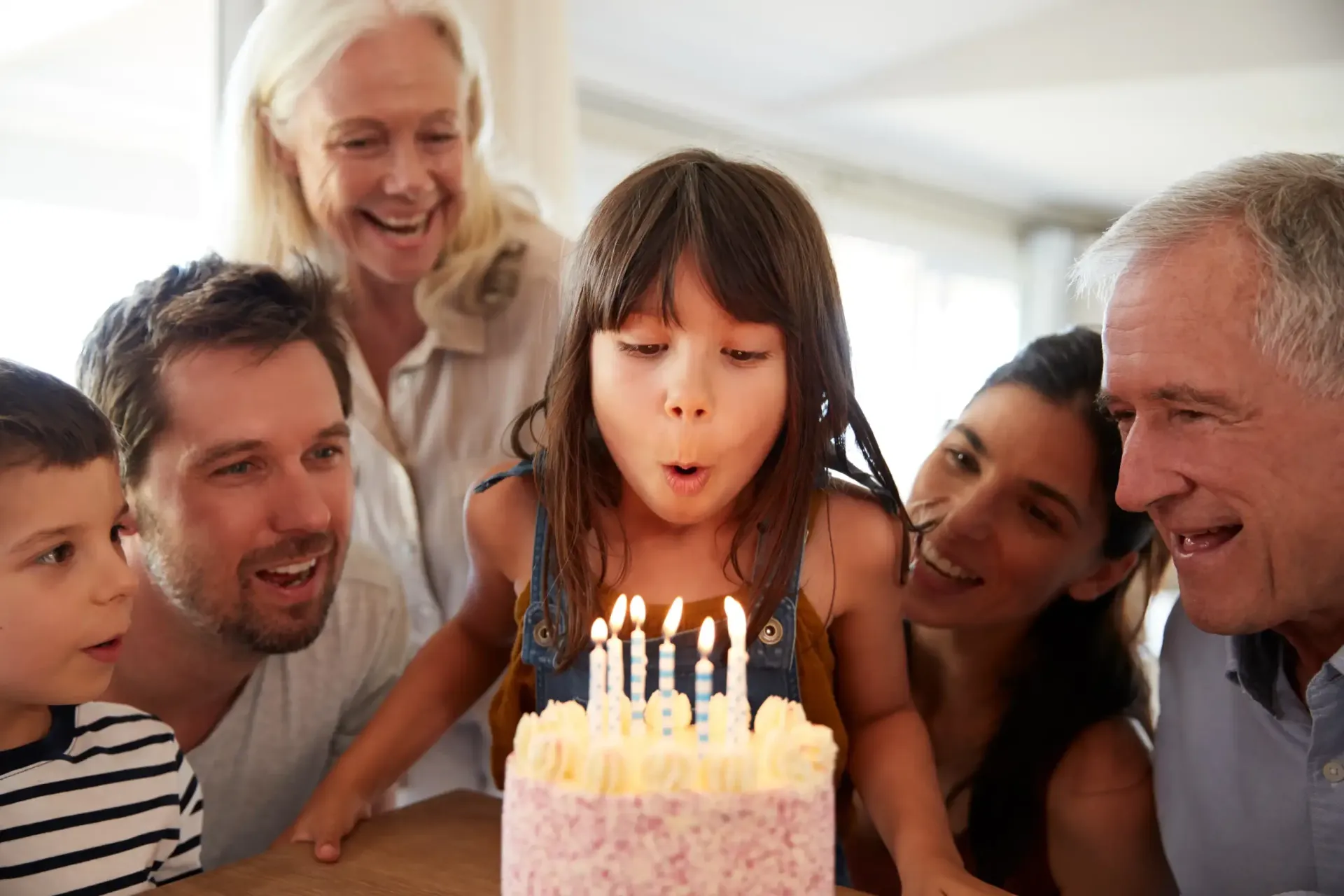 Girl blowing out candles on a birthday cake, surrounded by family, indoors, smiling.