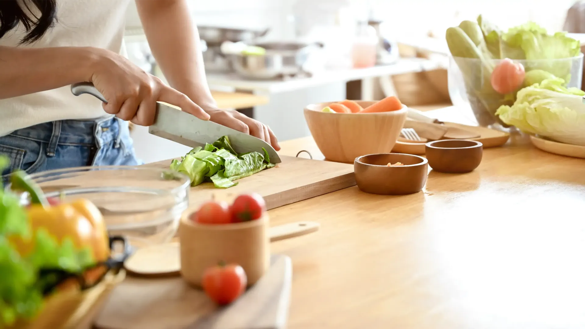 Person chopping lettuce on a wooden cutting board in a sunlit kitchen with vegetables in bowls.