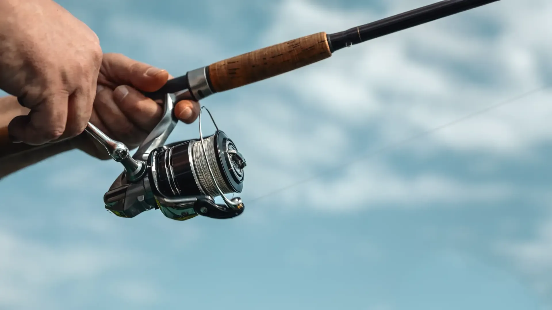 Person's hands holding a fishing rod and reel, with line visible, against a cloudy blue sky.