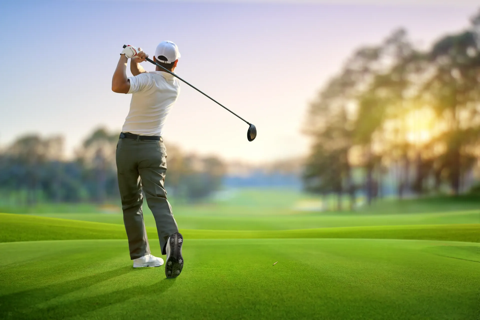 Golfer swinging a club on a green golf course, sunlight in the background.