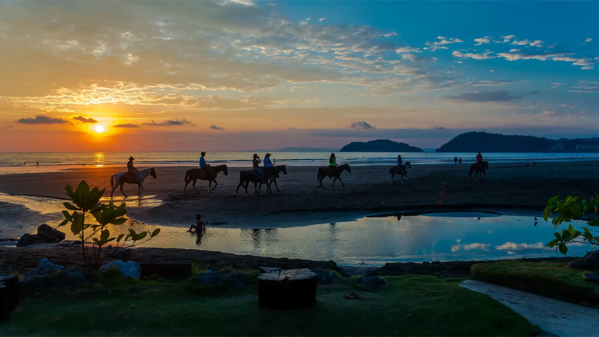 Horses and riders on a beach at sunset, with ocean, sky, and island in the distance.