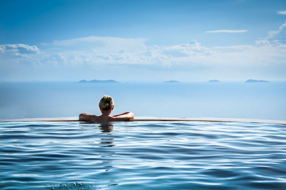 Woman in a pool, arms on edge, looking at ocean and islands under a bright blue sky.