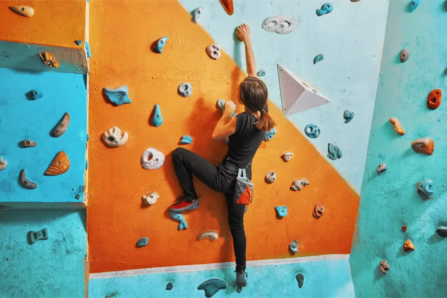 Person climbing an indoor rock wall, reaching for a hold. Orange and teal wall with various handholds.