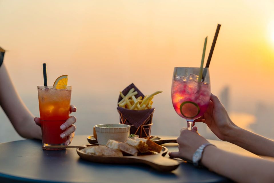 Two cocktails, fries, and appetizers on a table at sunset. Hands holding drinks.