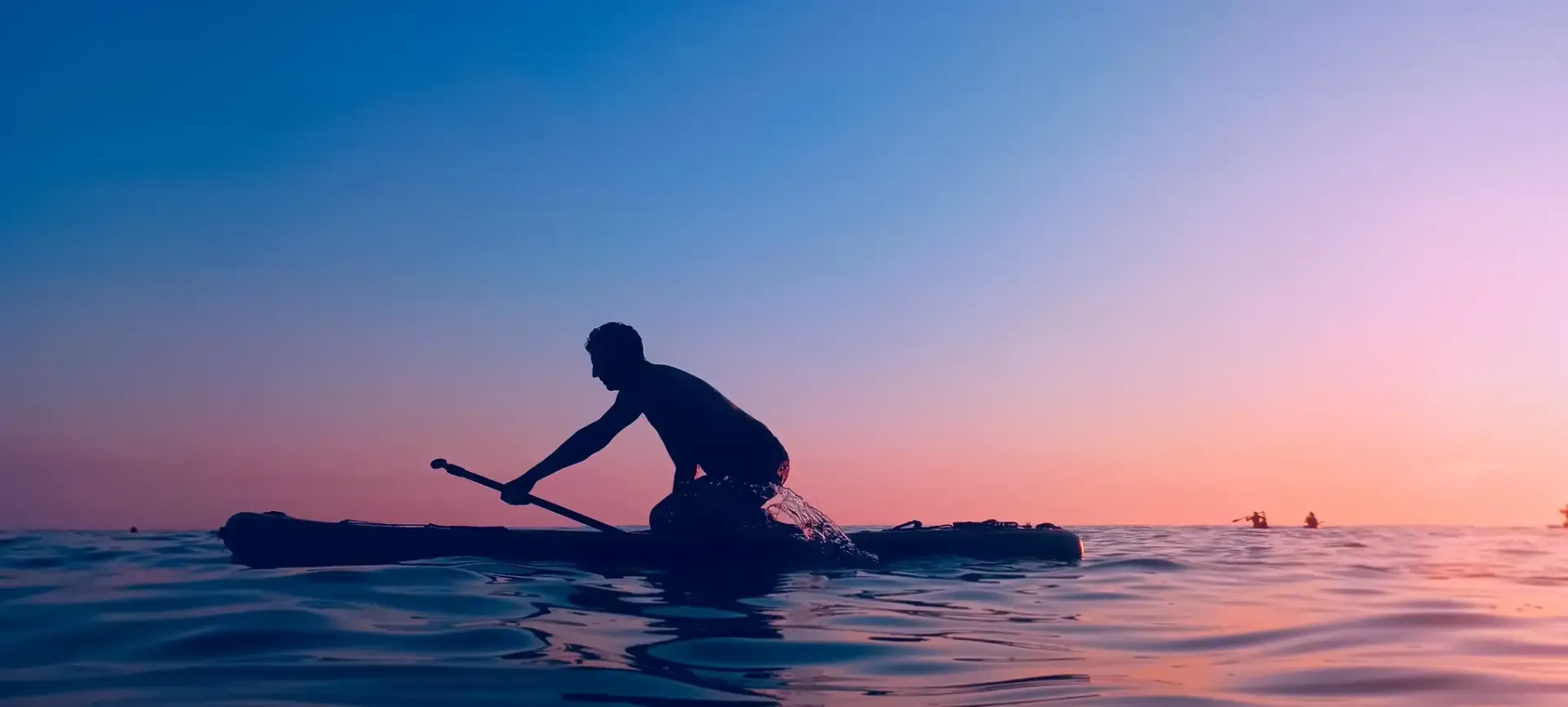 Silhouette of a person paddleboarding at sunset on calm water, with a blue to pink gradient sky.