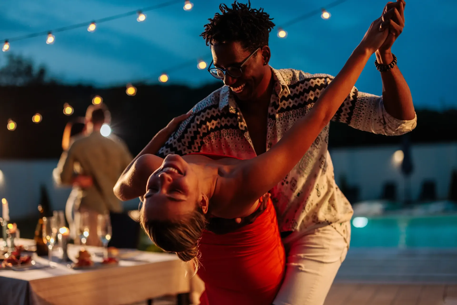 Couple dancing outdoors at night, smiling. Party with string lights, table, and pool.