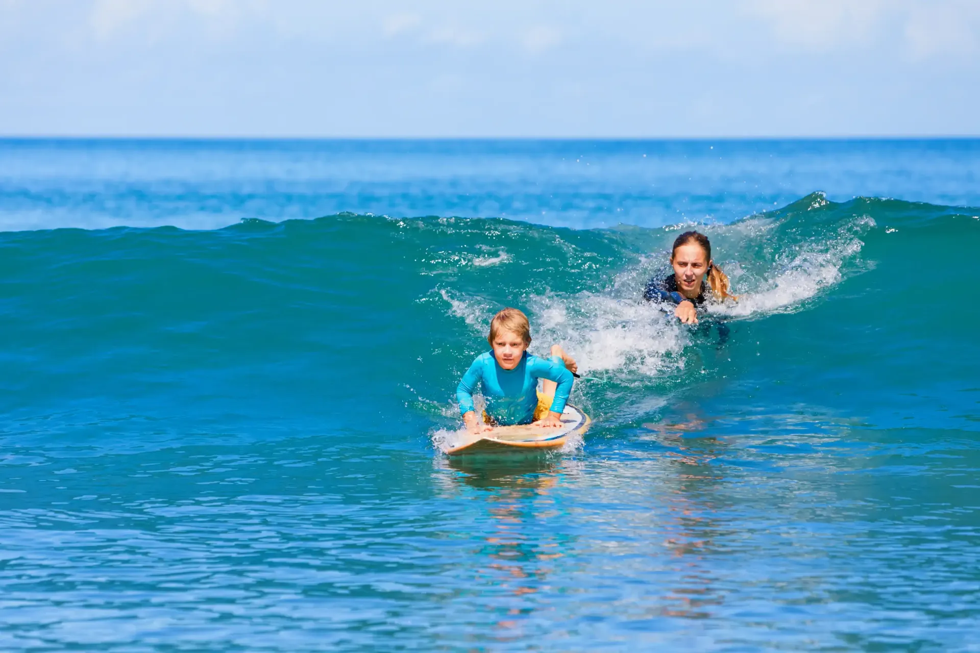 Person and child on surfboards in blue ocean water.