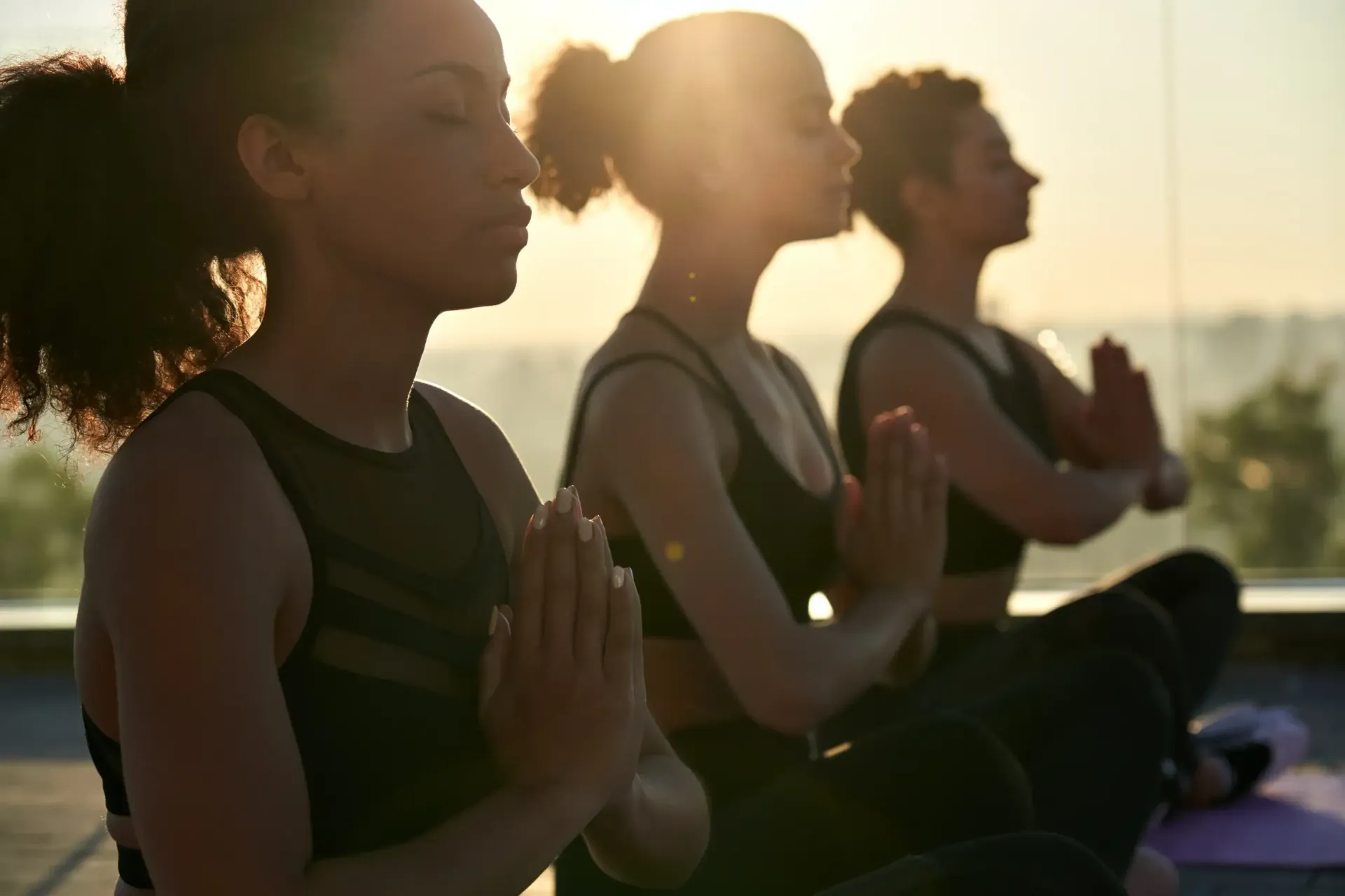 Three people in black activewear meditate with hands in prayer pose, outdoors with sunlight.